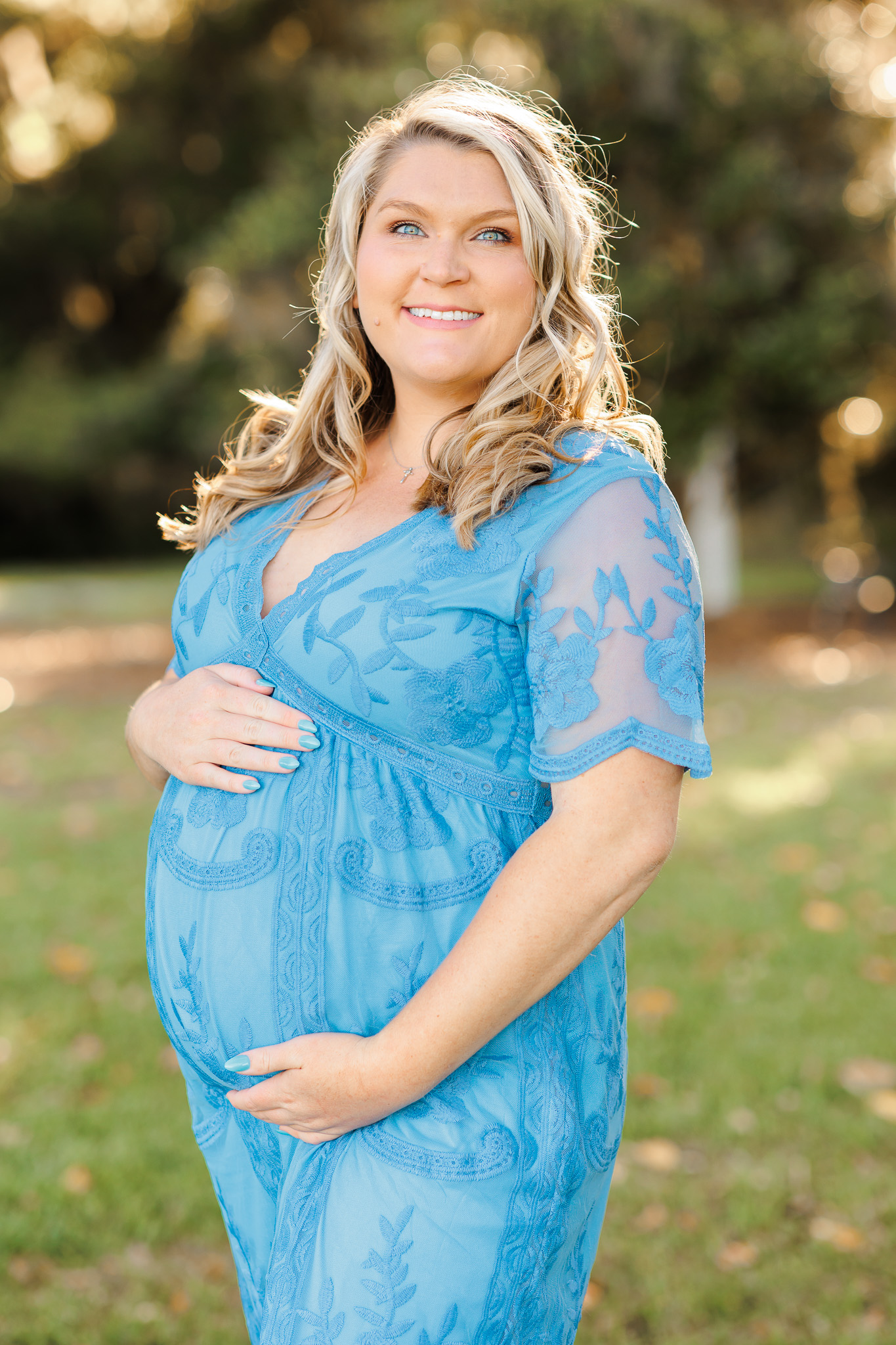 A pregnant woman in a blue lace dress holds her bump in a park