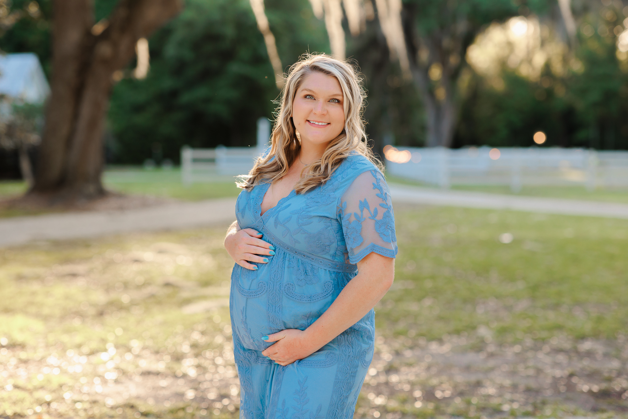 A mom-to-be stands at Hollyoak on the Marsh in a blue lace maternity gown with hands on her bump doulas in savannah ga