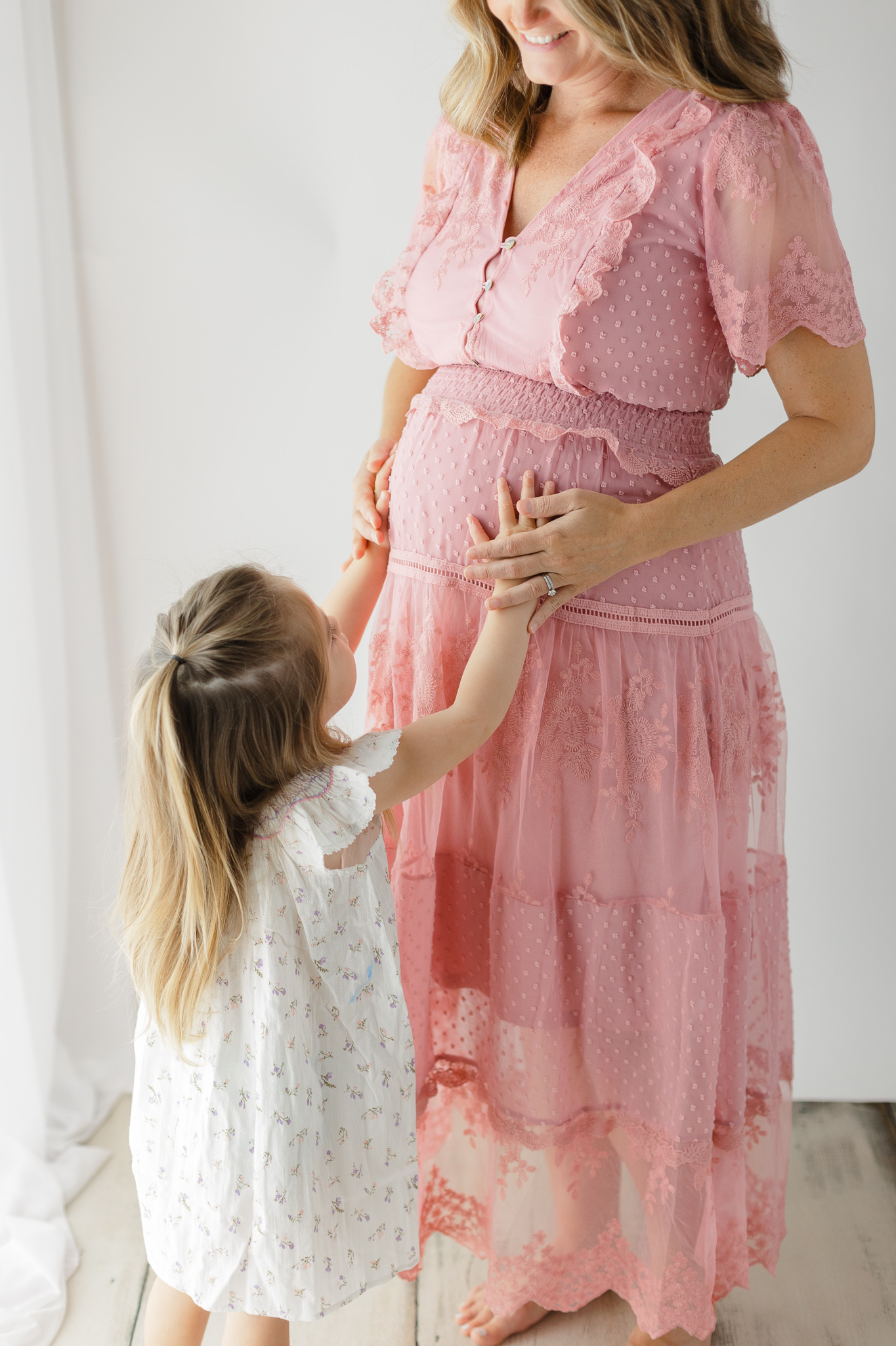 A pregnant mother stands by a window with her toddler toddler reaching up to feel her bump