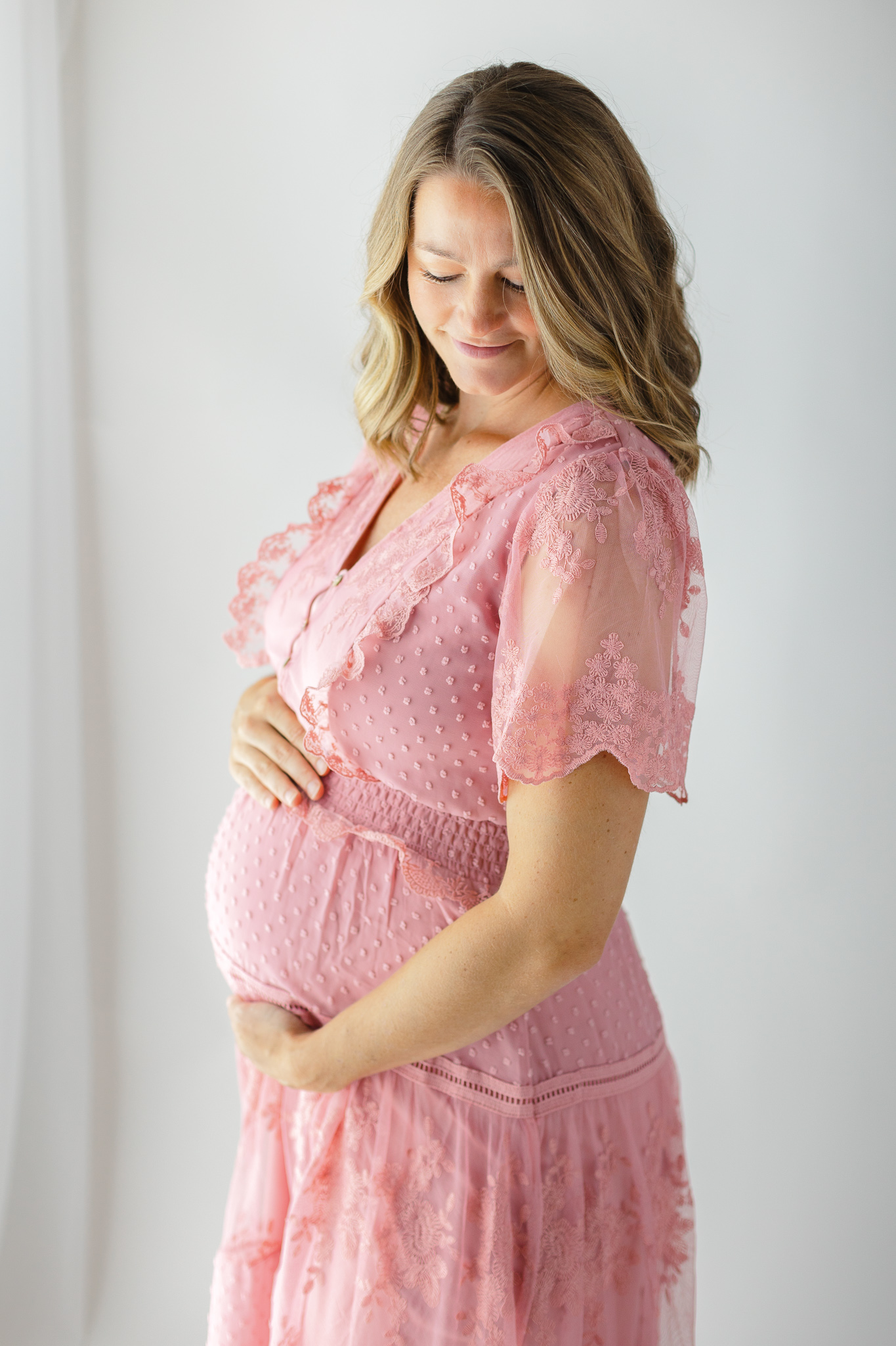 A mother to be looks down her shoulder while standing by a window in a pink maternity dress midwives savannah ga