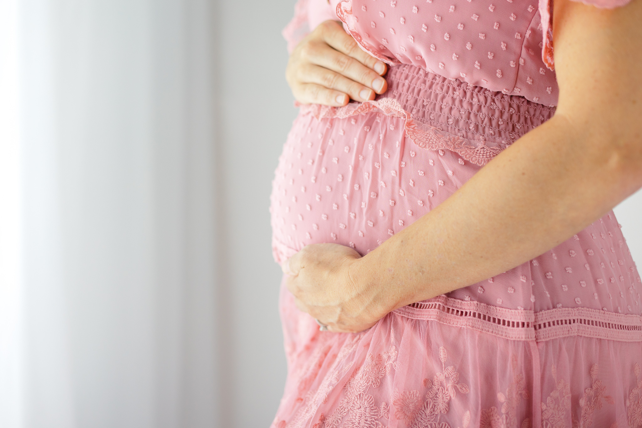 A mom-to-be stands in front of a tall window in a pink dress with hands on her bump midwives savannah ga