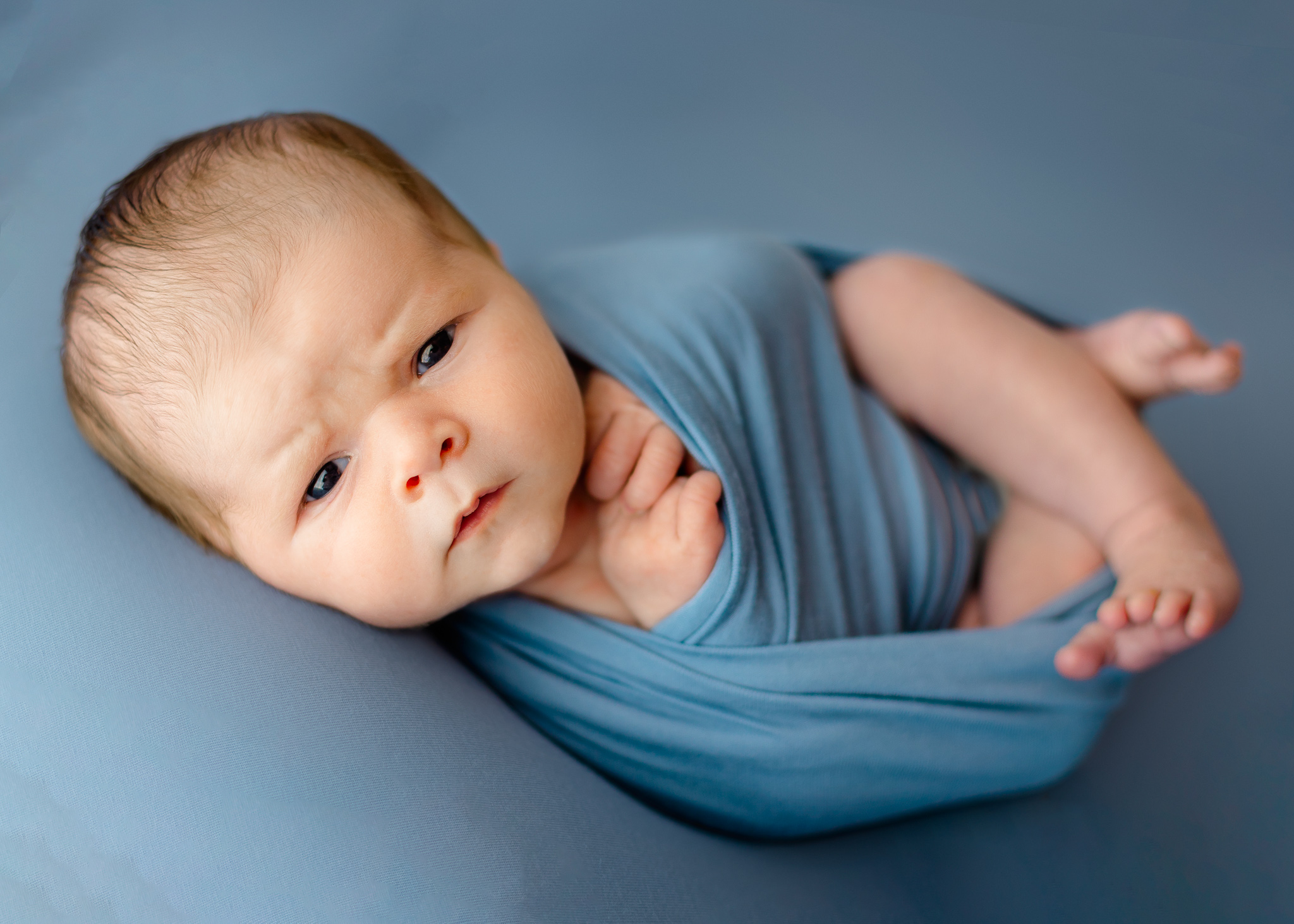 A newborn baby lays in a blue swaddle with legs sticking out and eyes open on a blue bed