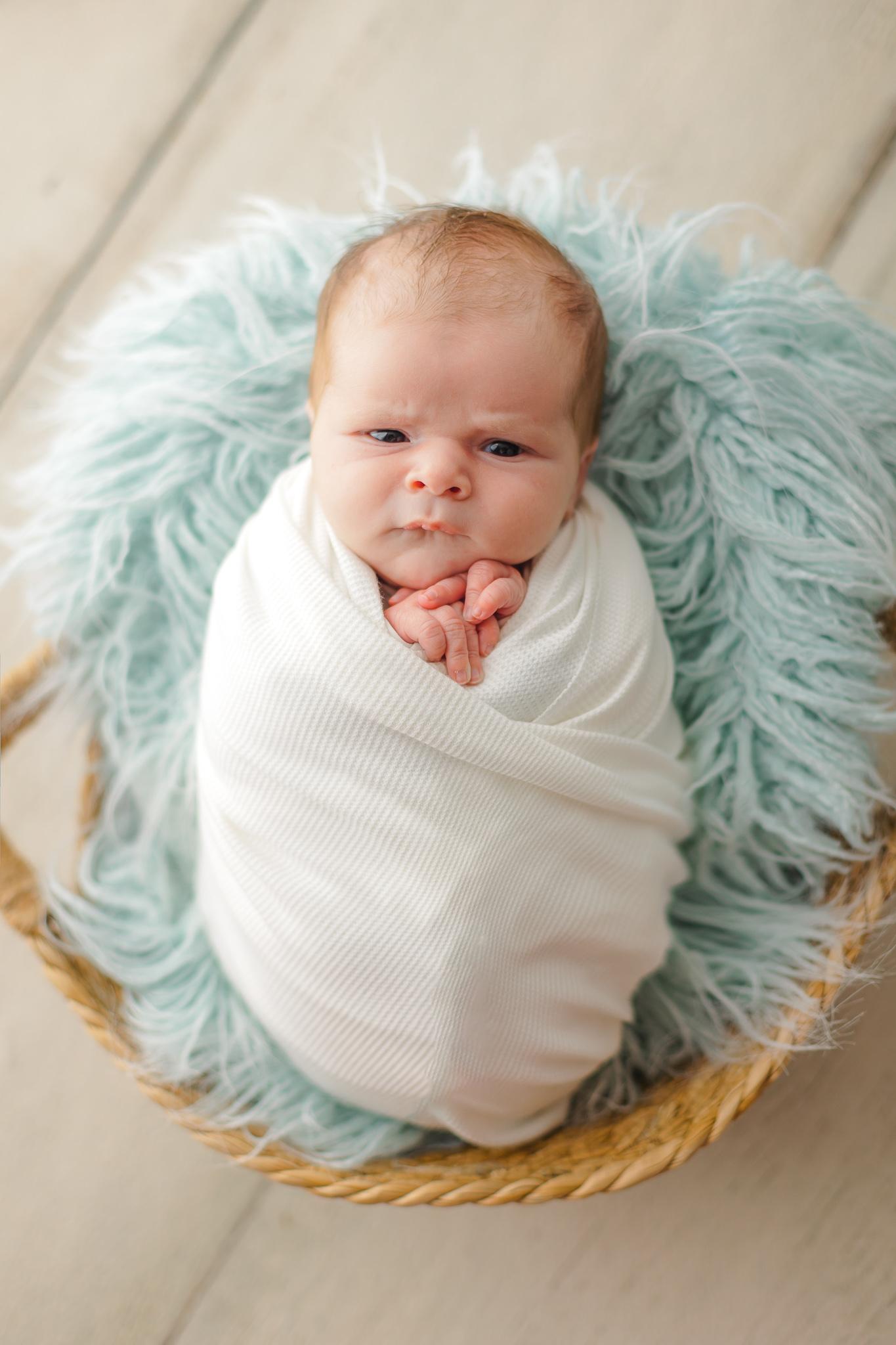 A sleepy newborn baby in a white swaddle lays on a blue fur blanket in a woven basket savannah baby stores