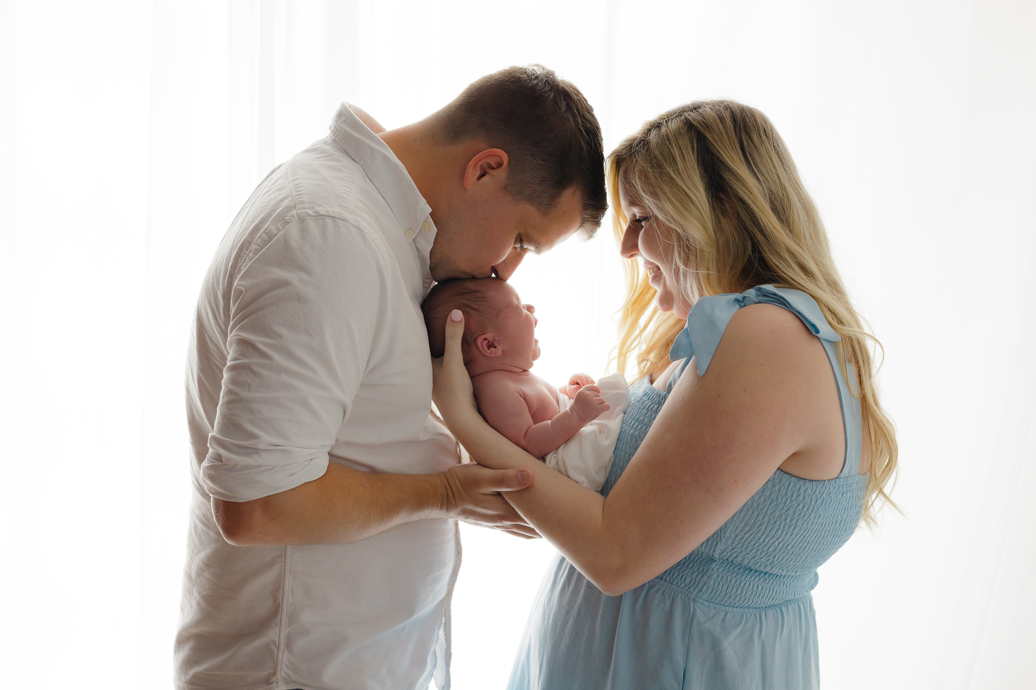 A father kisses the forehead of his crying newborn baby that mom is holding in front of a window