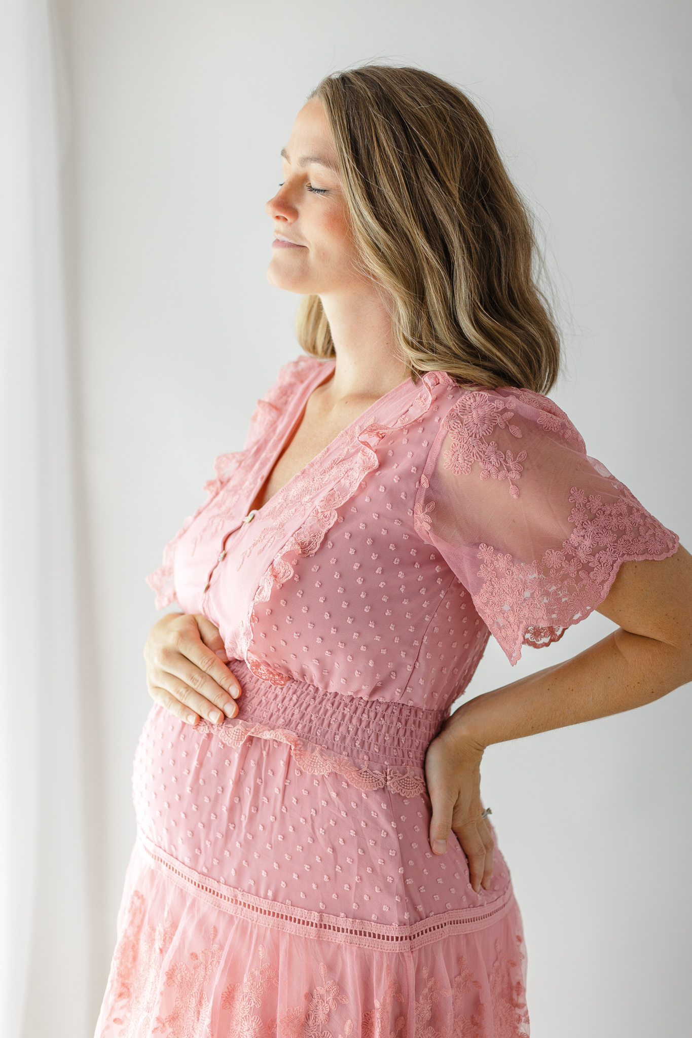 A mom to be stands by a window soaking in the sunlight in a pink maternity dress prenatal massage Savannah