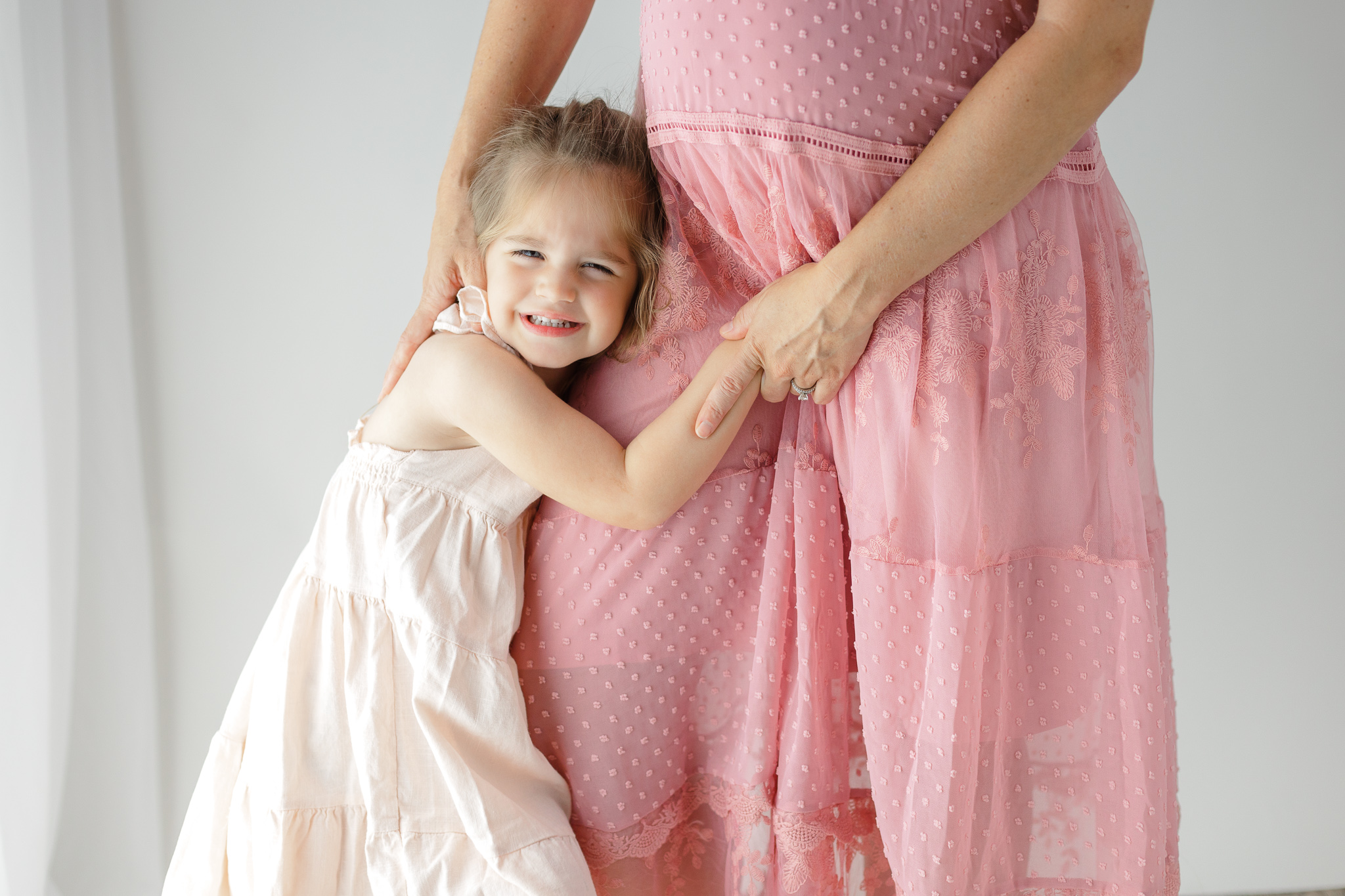 A young girl in a white dress hugs the leg of her pregnant mother