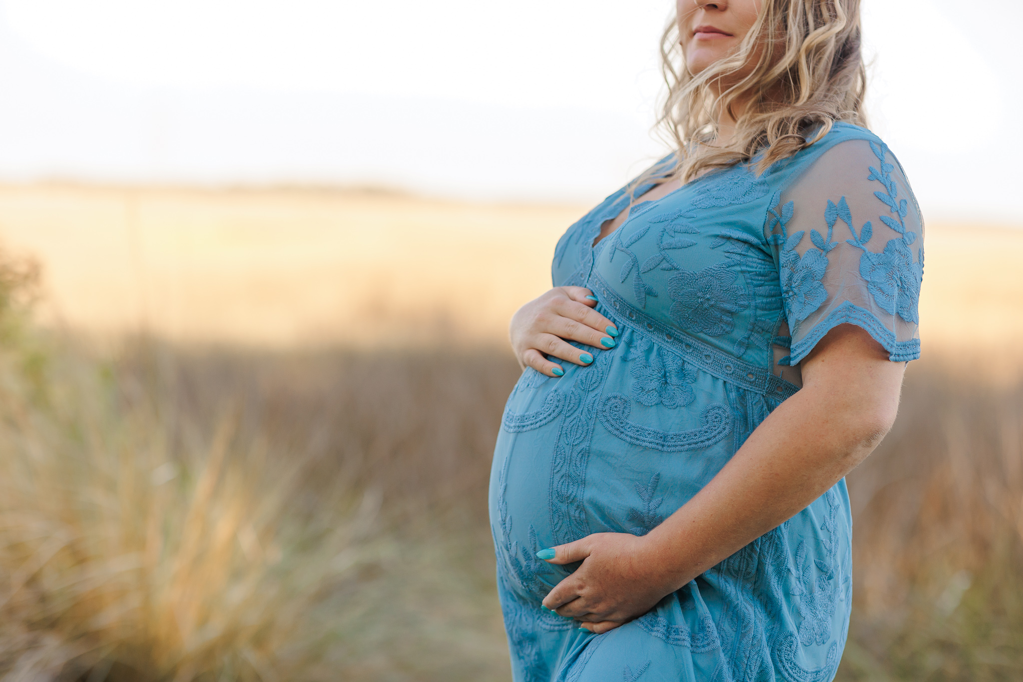 A mother to be holds her bump in a golden grass field while wearing a blue lace maternity gown