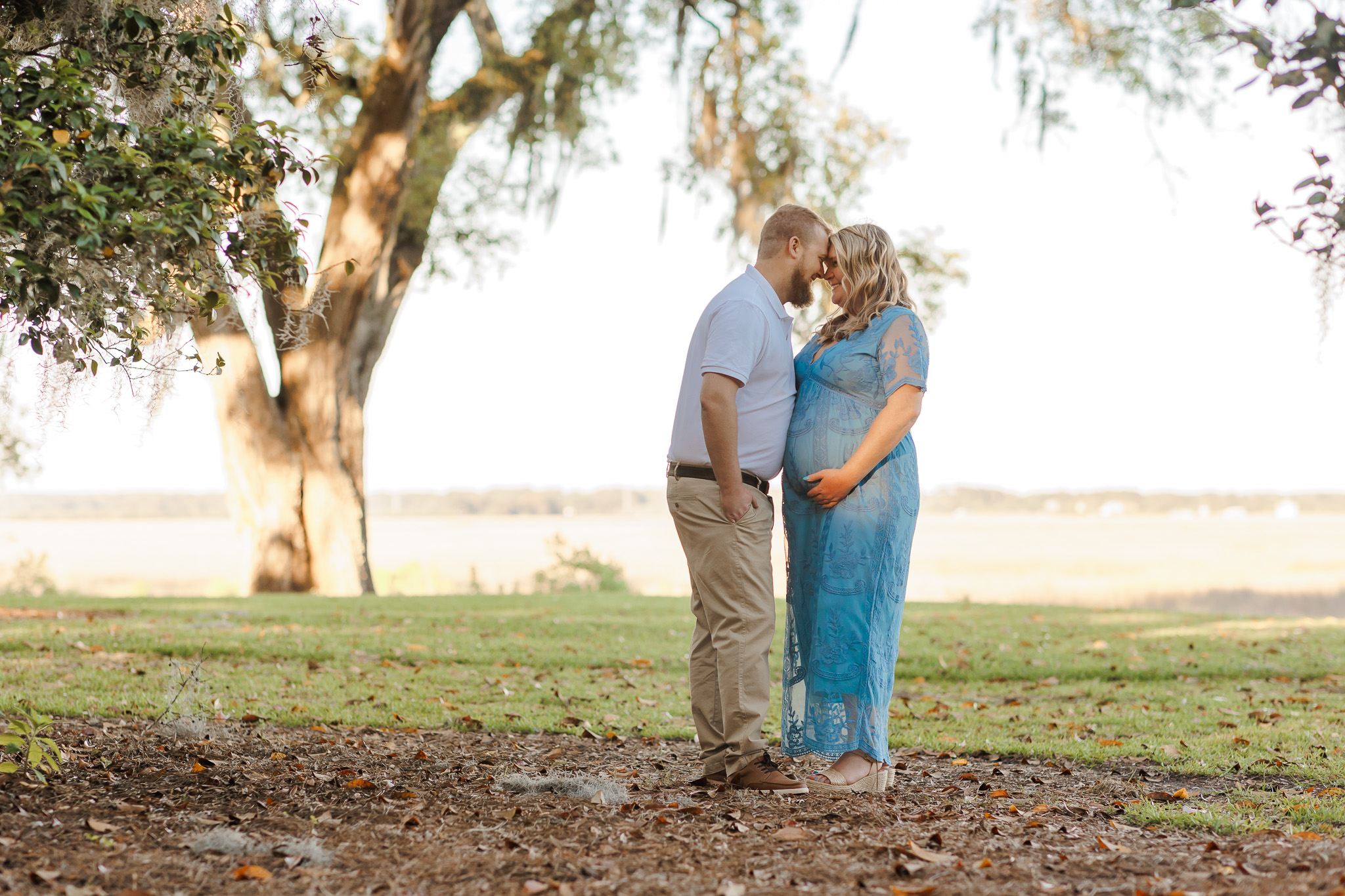 A mother to be stands in a park in a blue maternity gown placing her forehead to her husbands prenatal yoga Savannah