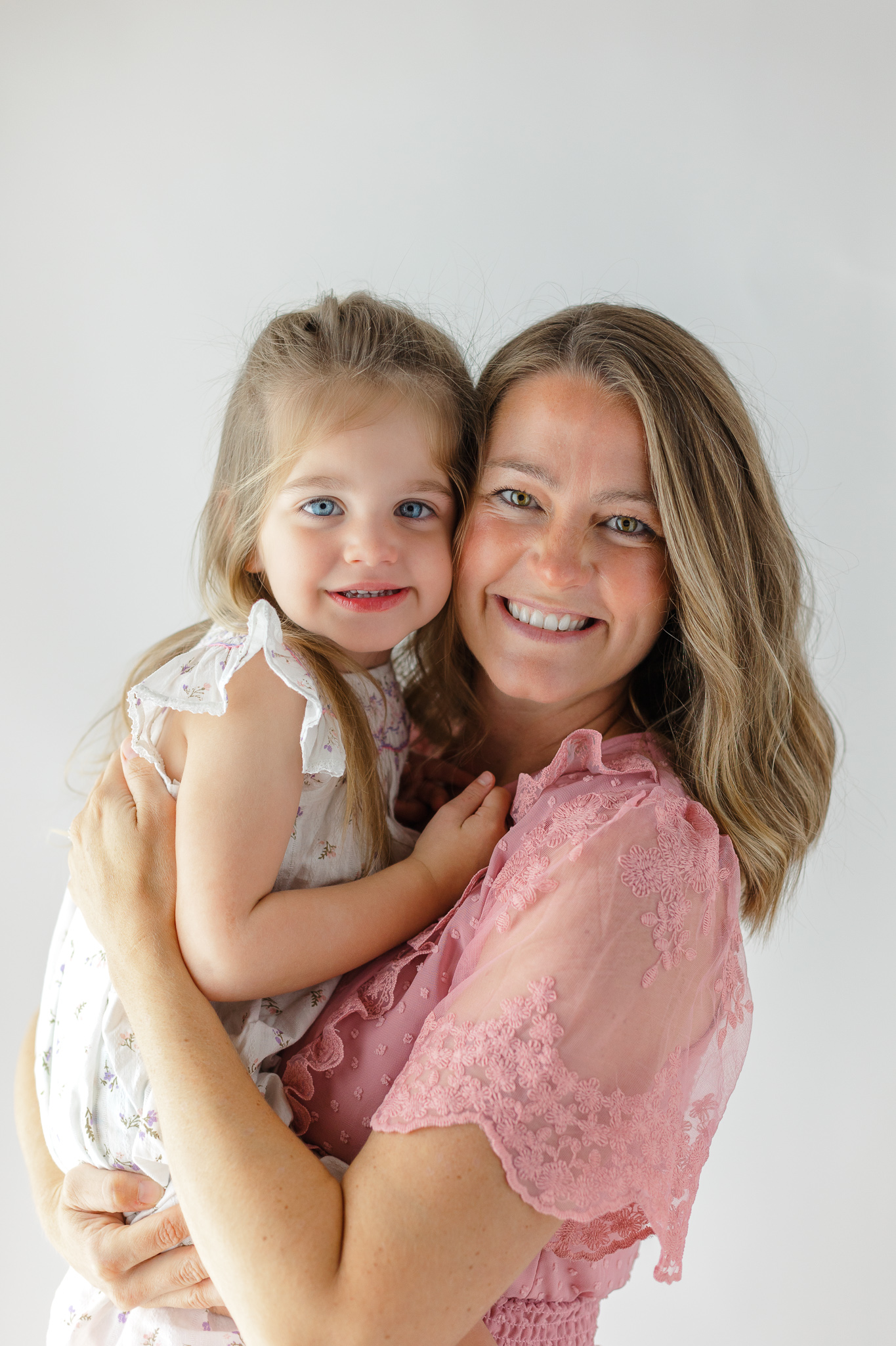 A mom in a pink dress holds her toddler daughter cheek to cheek in a studio running out of womb