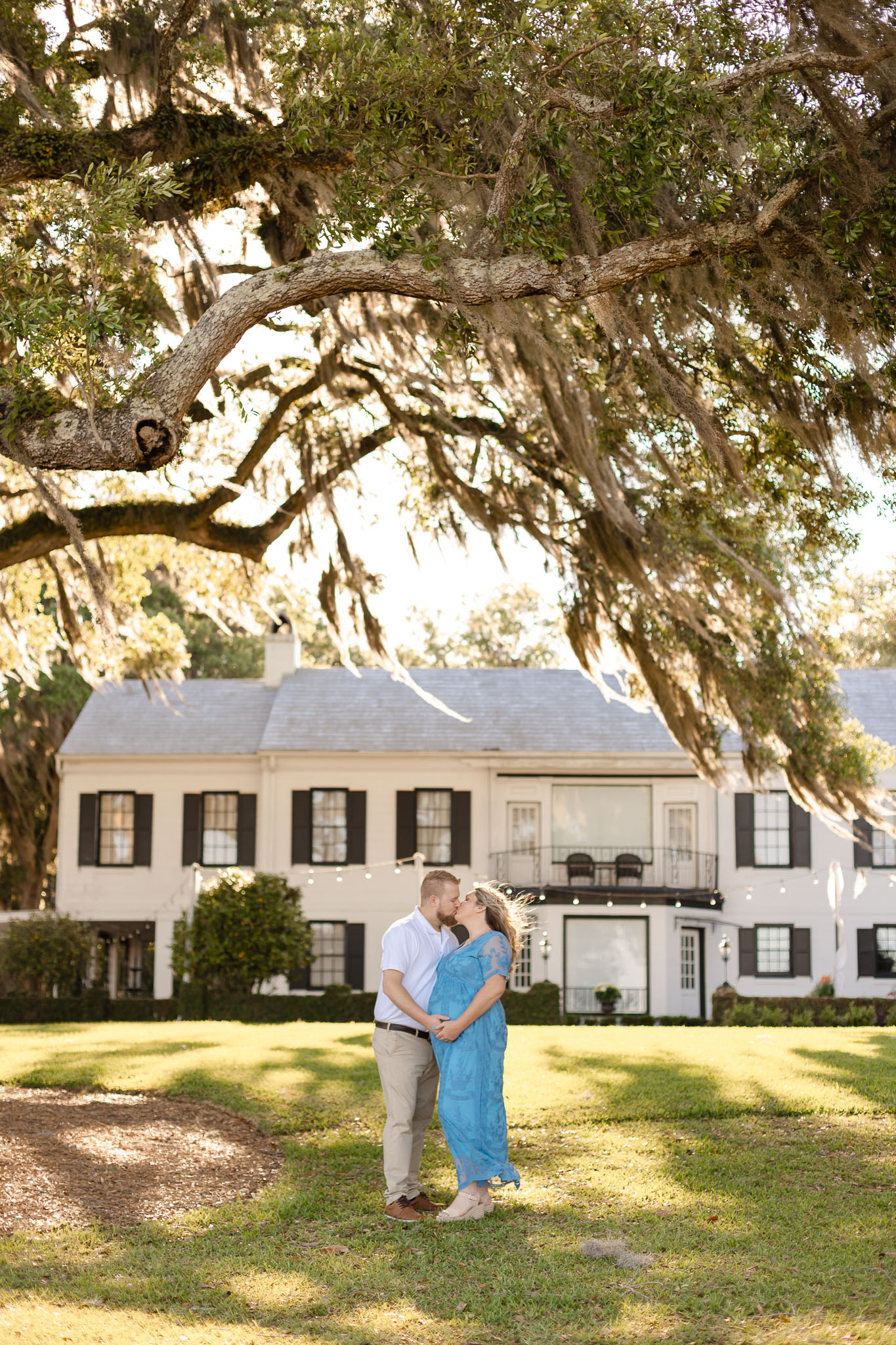 A couple kiss under an oak tree while holding the pregnant bump of the woman in a blue lace dress savannah fertility clinic