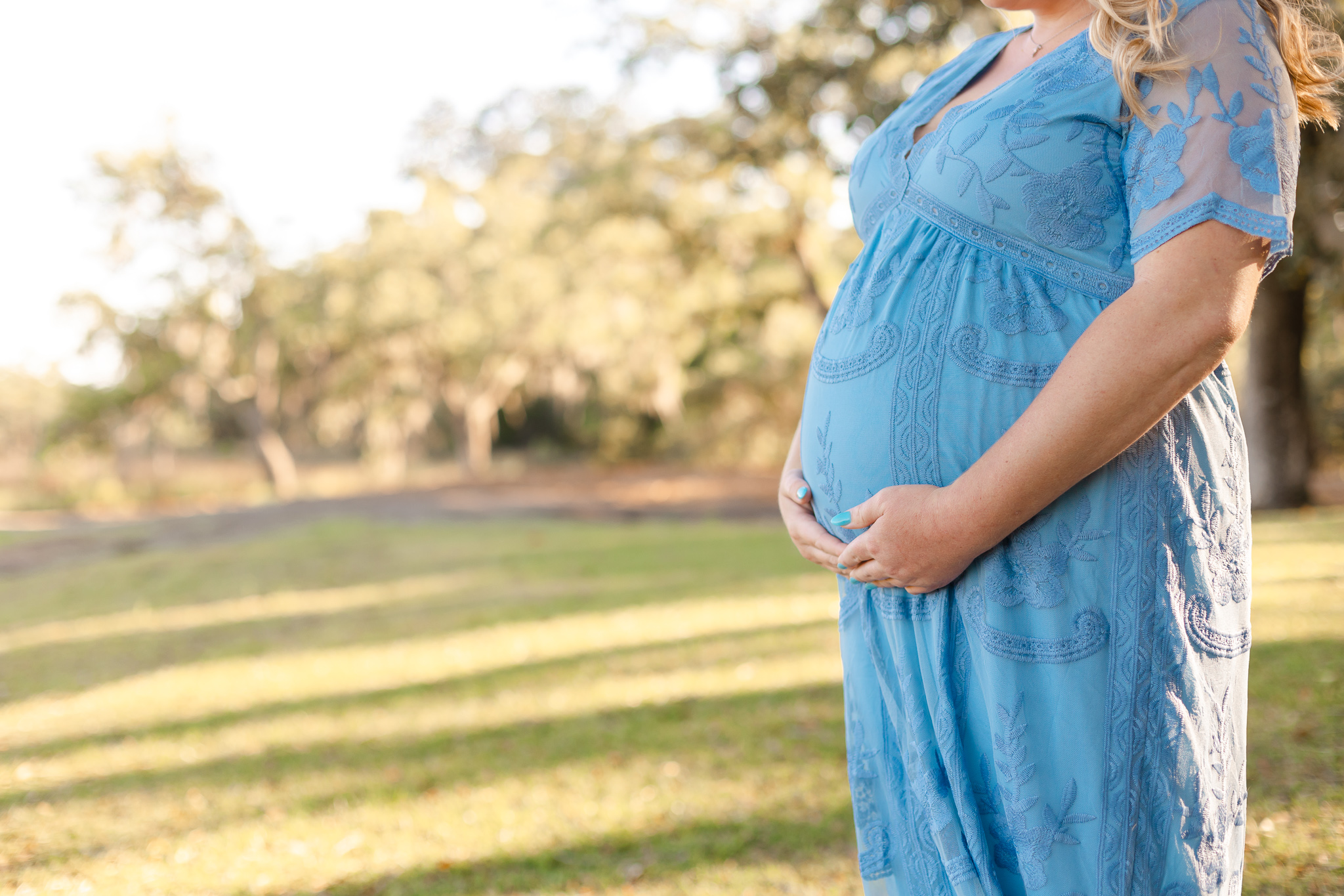 A mother to be stands in a blue lace maternity gown in a park field savannah fertility clinic