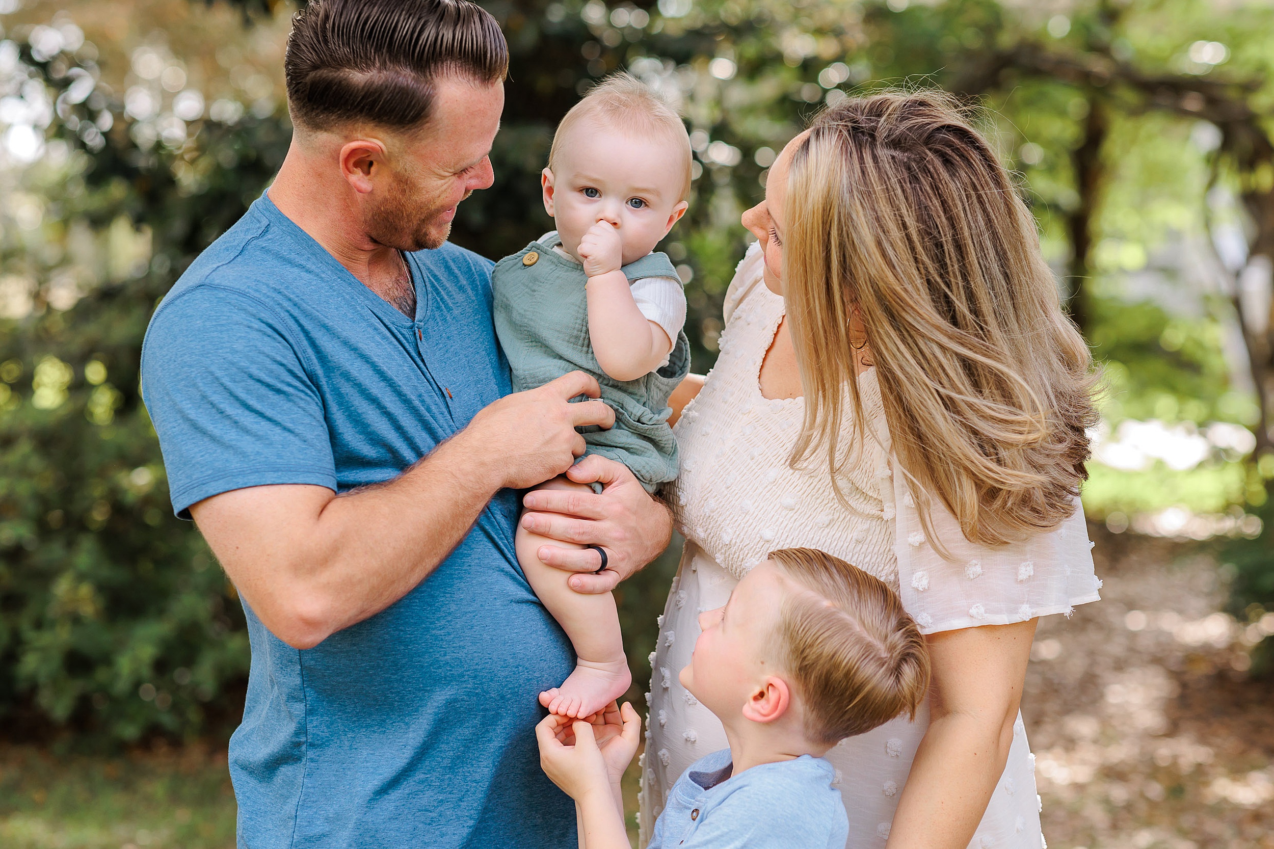 A dad holds his infant son while mom and their younger son tickle the infant sucking his thumb