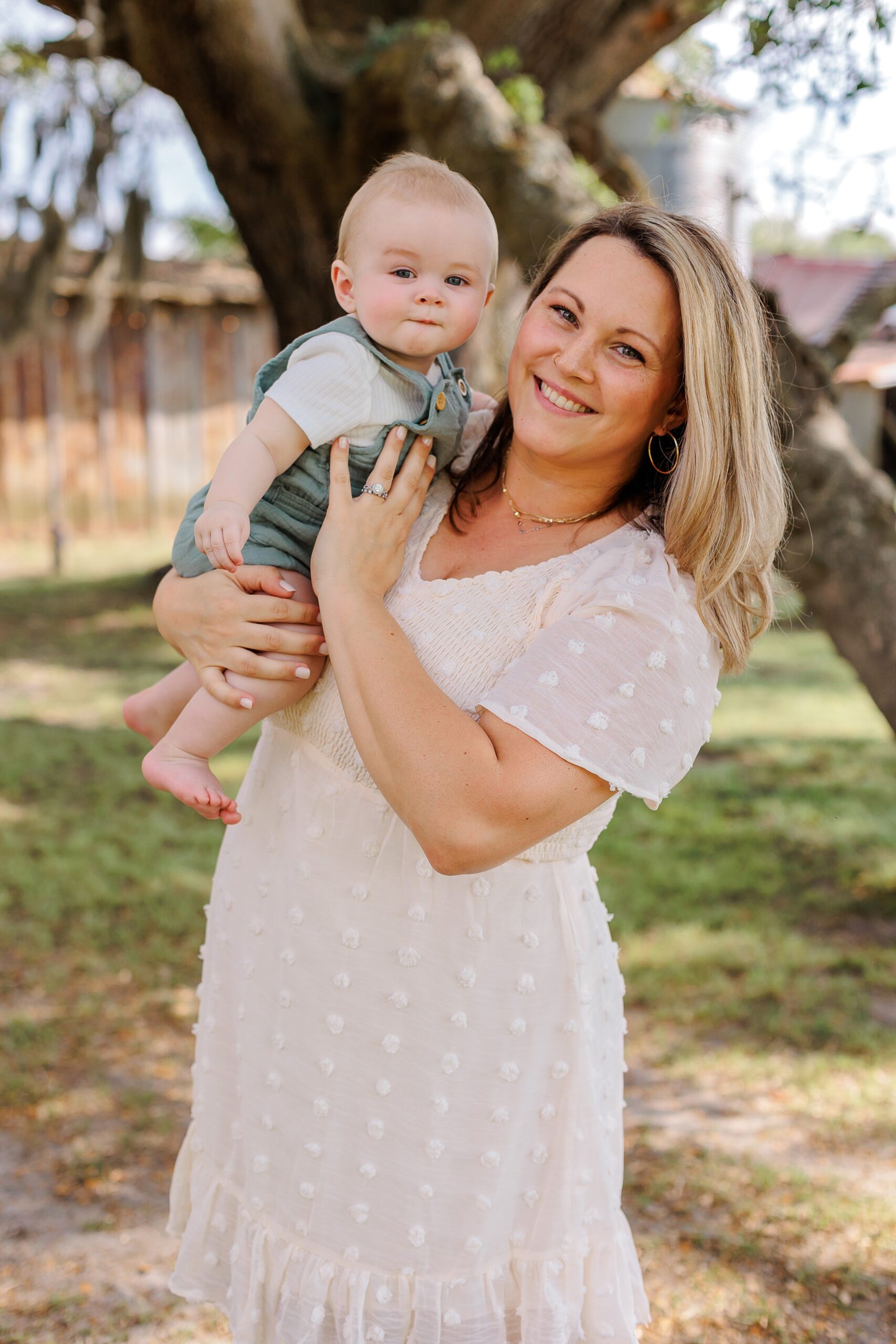 A mother in a white dress stands under a tree holding her infant son in green overalls nannies in Savannah