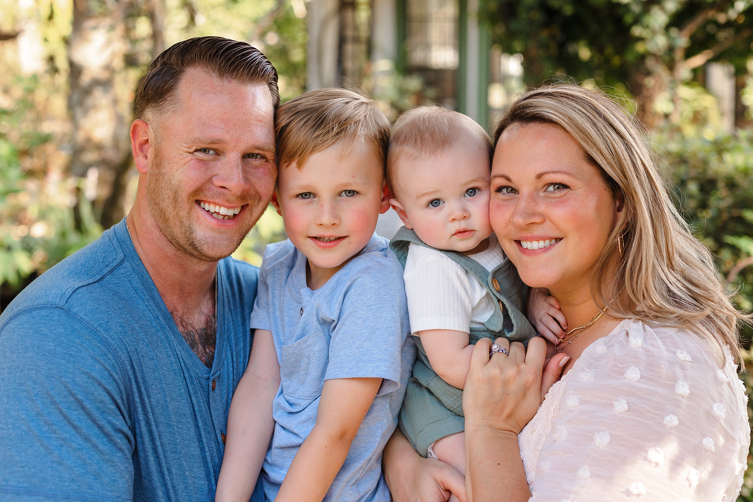 A mom and dad press their cheeks together with their two sons while standing in a garden