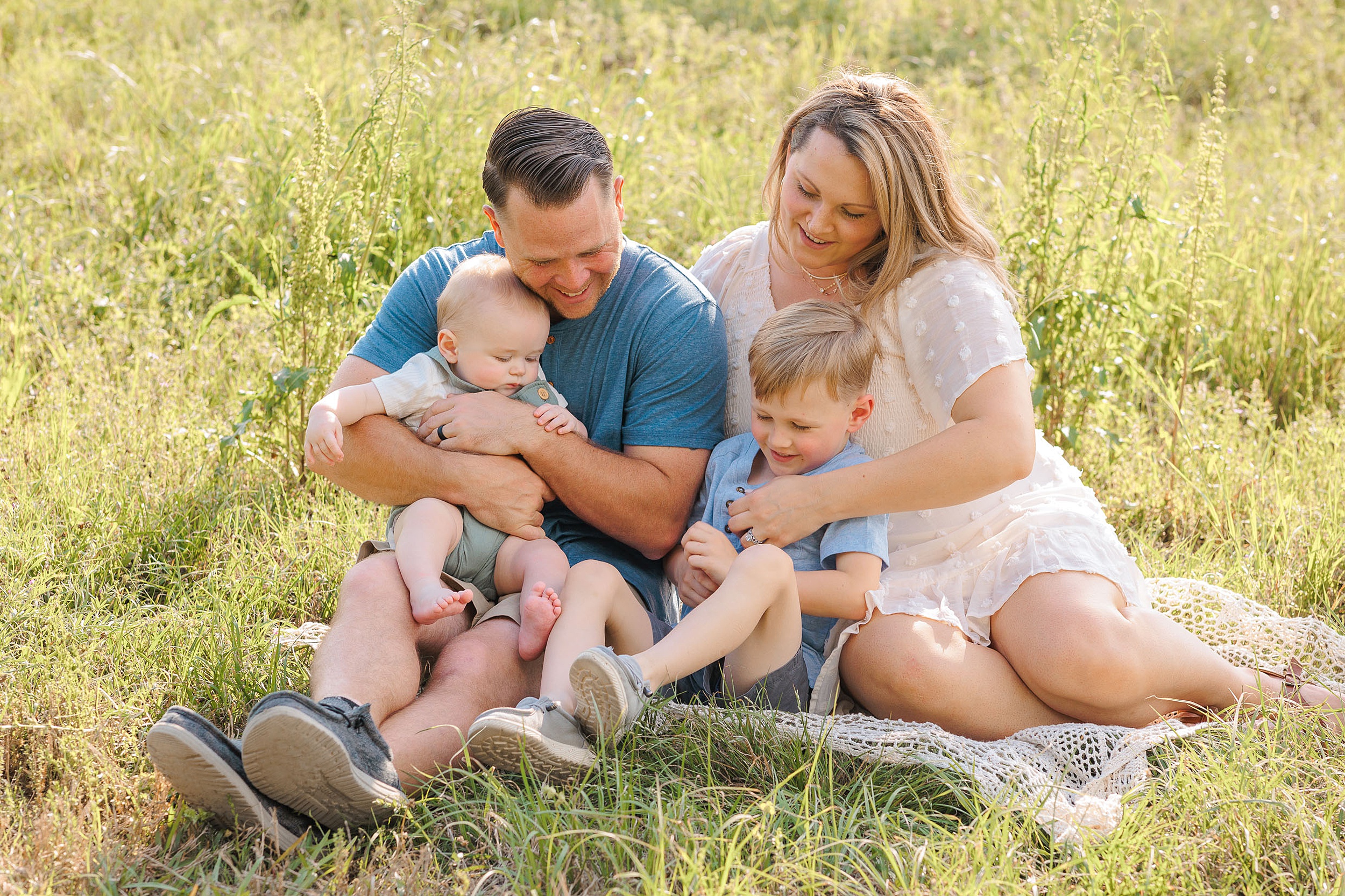 A mom and dad sit on a picnic blanket in a grassy field playing with their two young sons in their laps savannah pediatric dentist