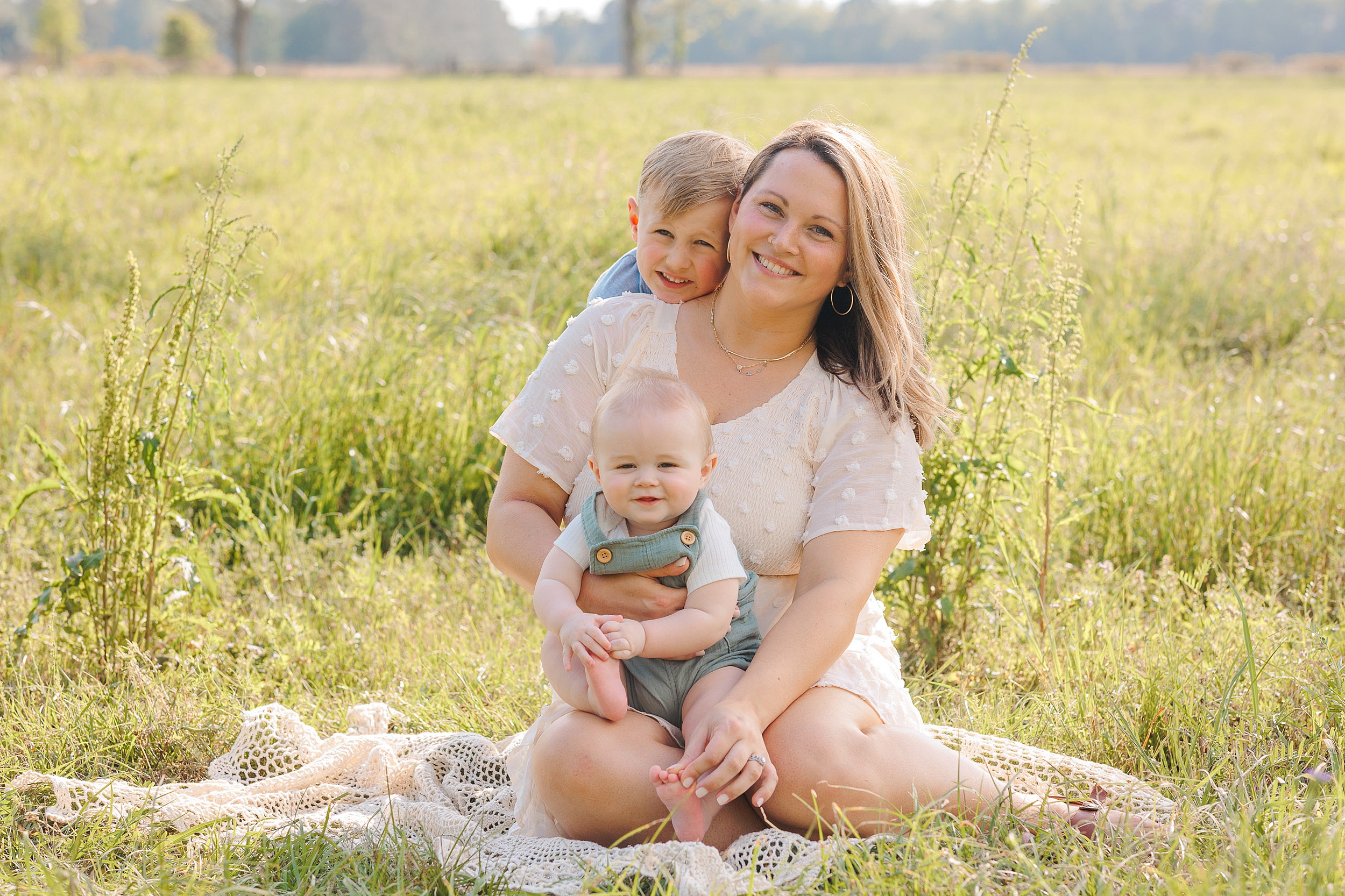 A mother in a white dress sits on a blanket in a field of grass with her infant in her lap and older son climbing on her back savannah pediatric dentist