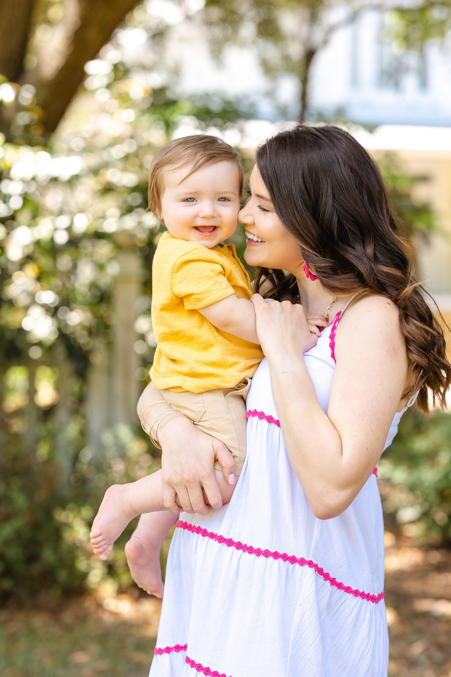 A mom in a white and pink dress snuggles and plays with her infant son on her hip