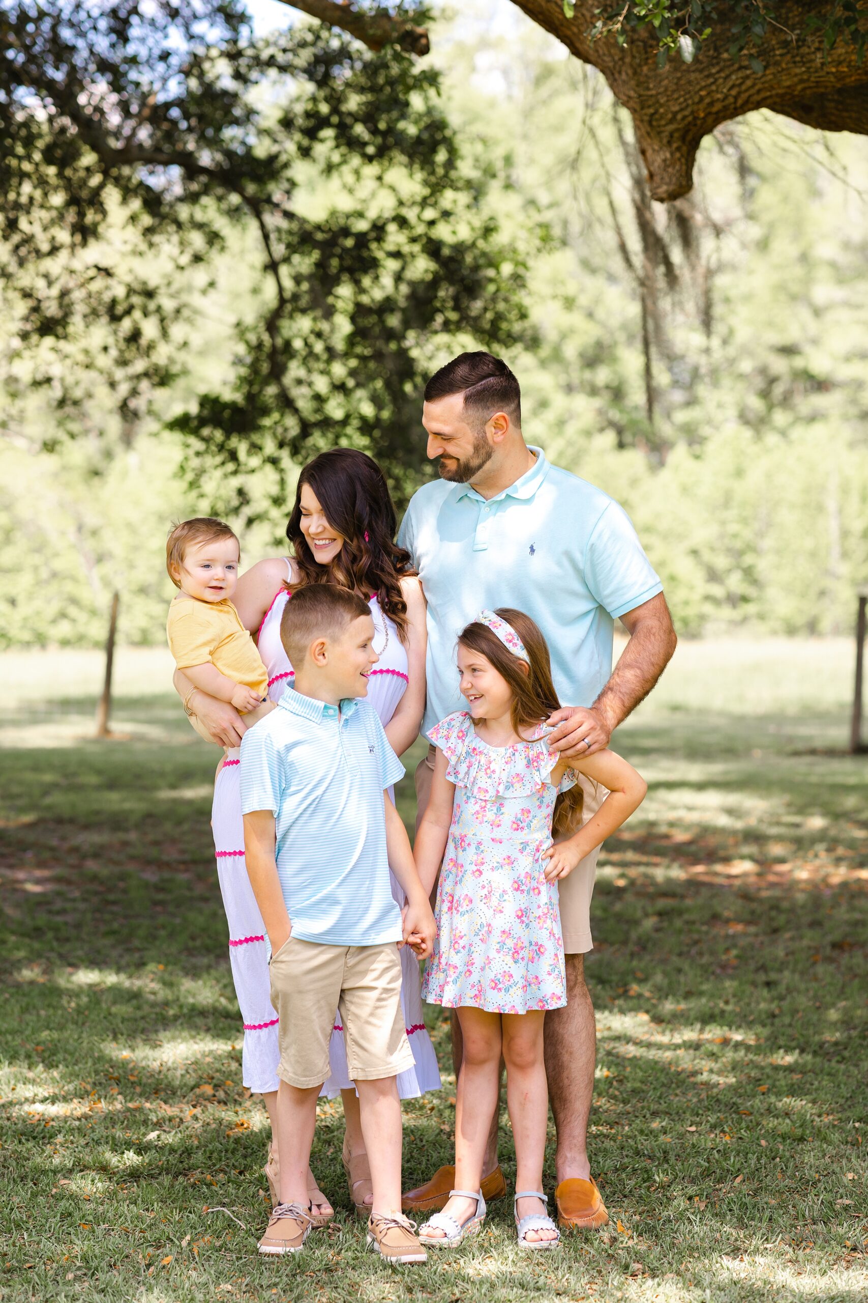 A family of five stand together in a park under a tree holding hands and looking at the infant on mom's hip savannah pediatricians