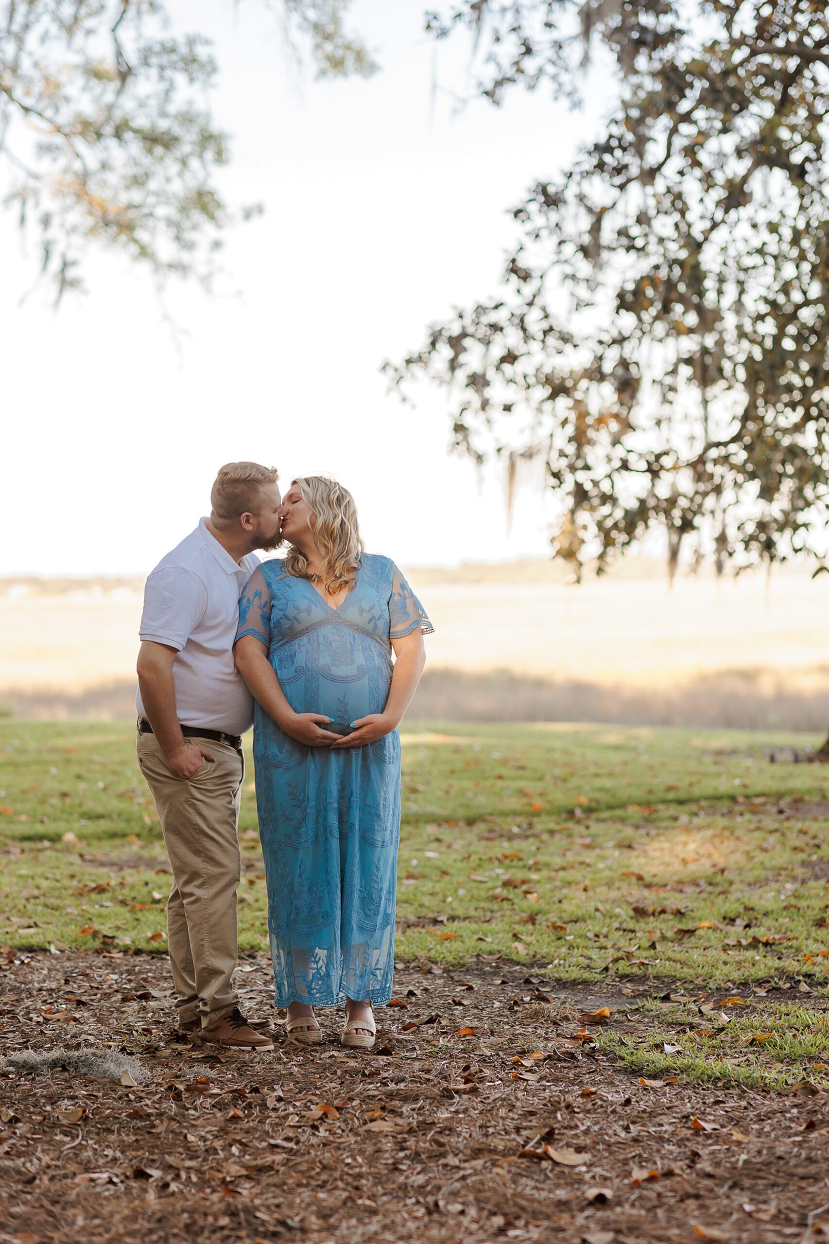 A mom to be stands holding her bump in a blue dress while kissing her husband in a white polo and khakis in a park