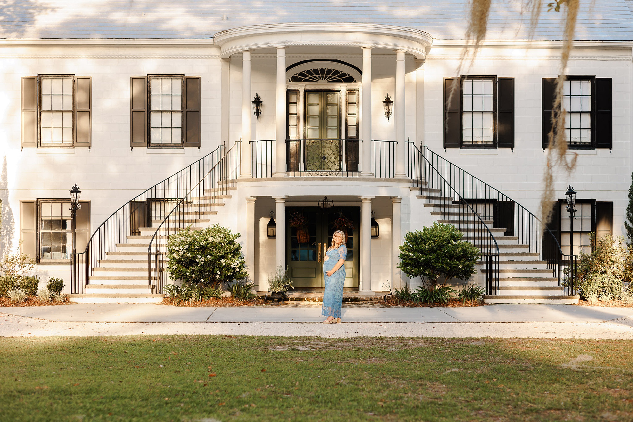 A mom to be in a blue lace dress holds her bump while standing in front of a two story entrance to a large white building with two staircases on a savannah babymoon