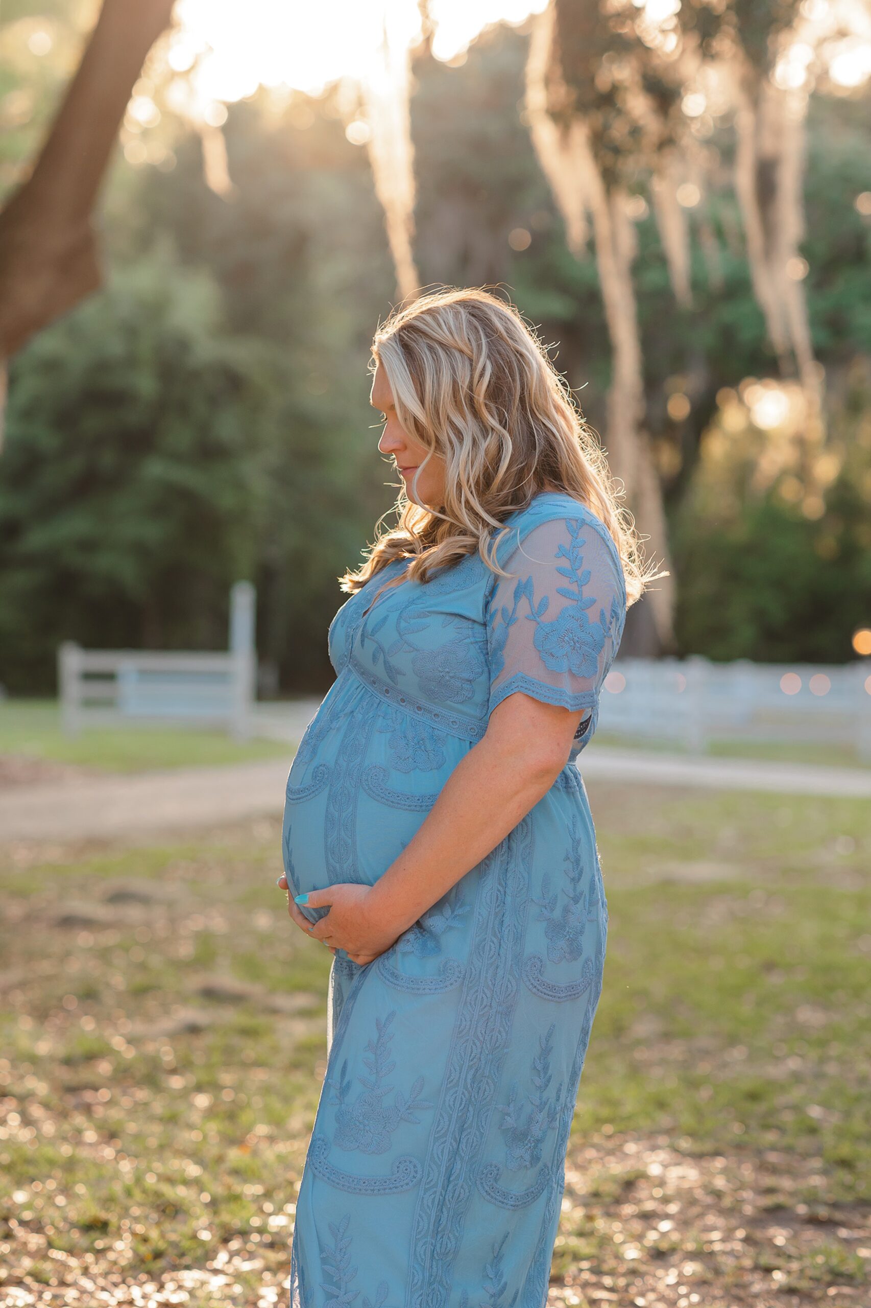 A mom to be in a blue lace dress stands under a tree in a park at sunset while looking down at her hands on her bump on a savannah babymoon