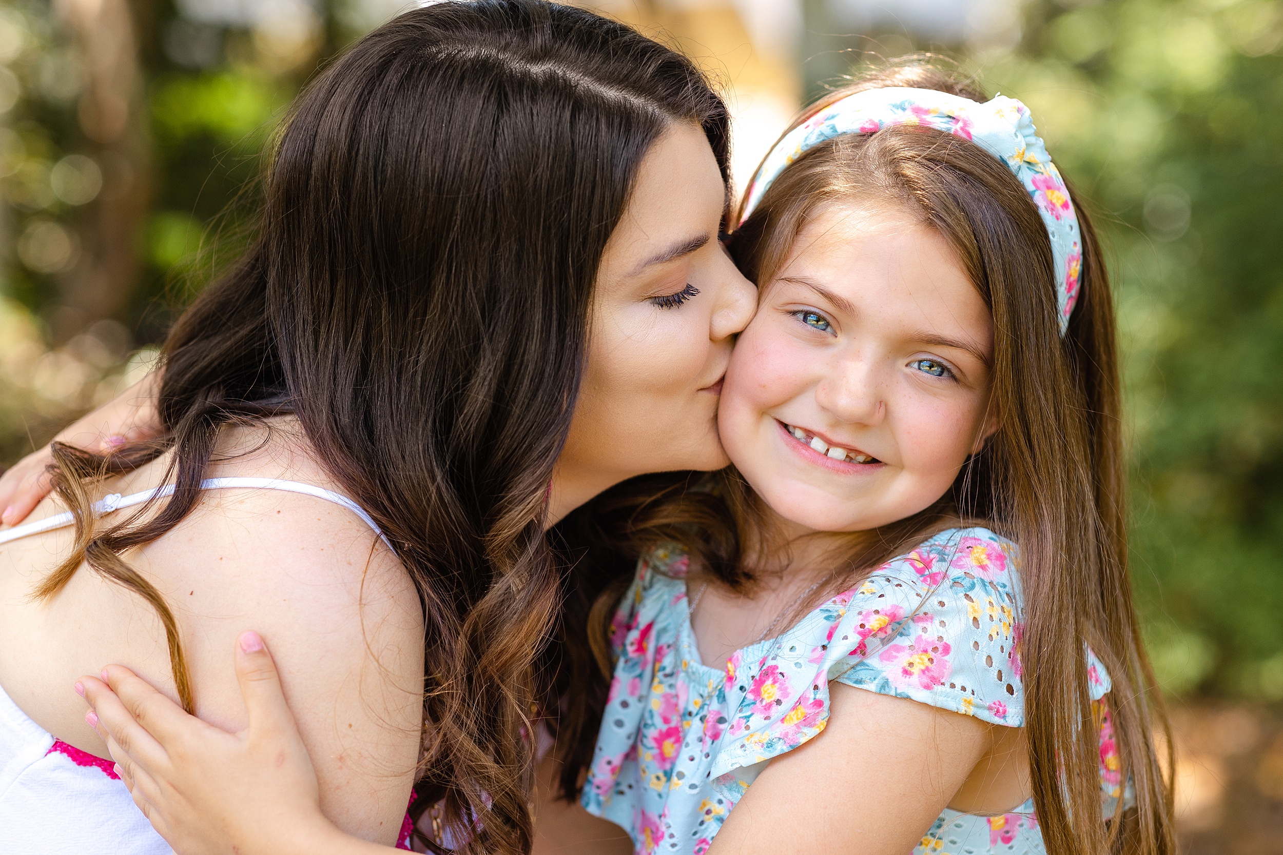 A mother with dark hair and a white dress kisses the cheek of her young daughter in a colorful floral dress and matching headband in savannah playgrounds