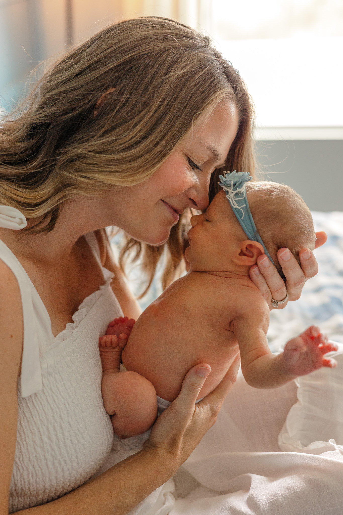 A mother nuzzles her nose to her newborn baby's forehead while they sit on a bed in front of a window