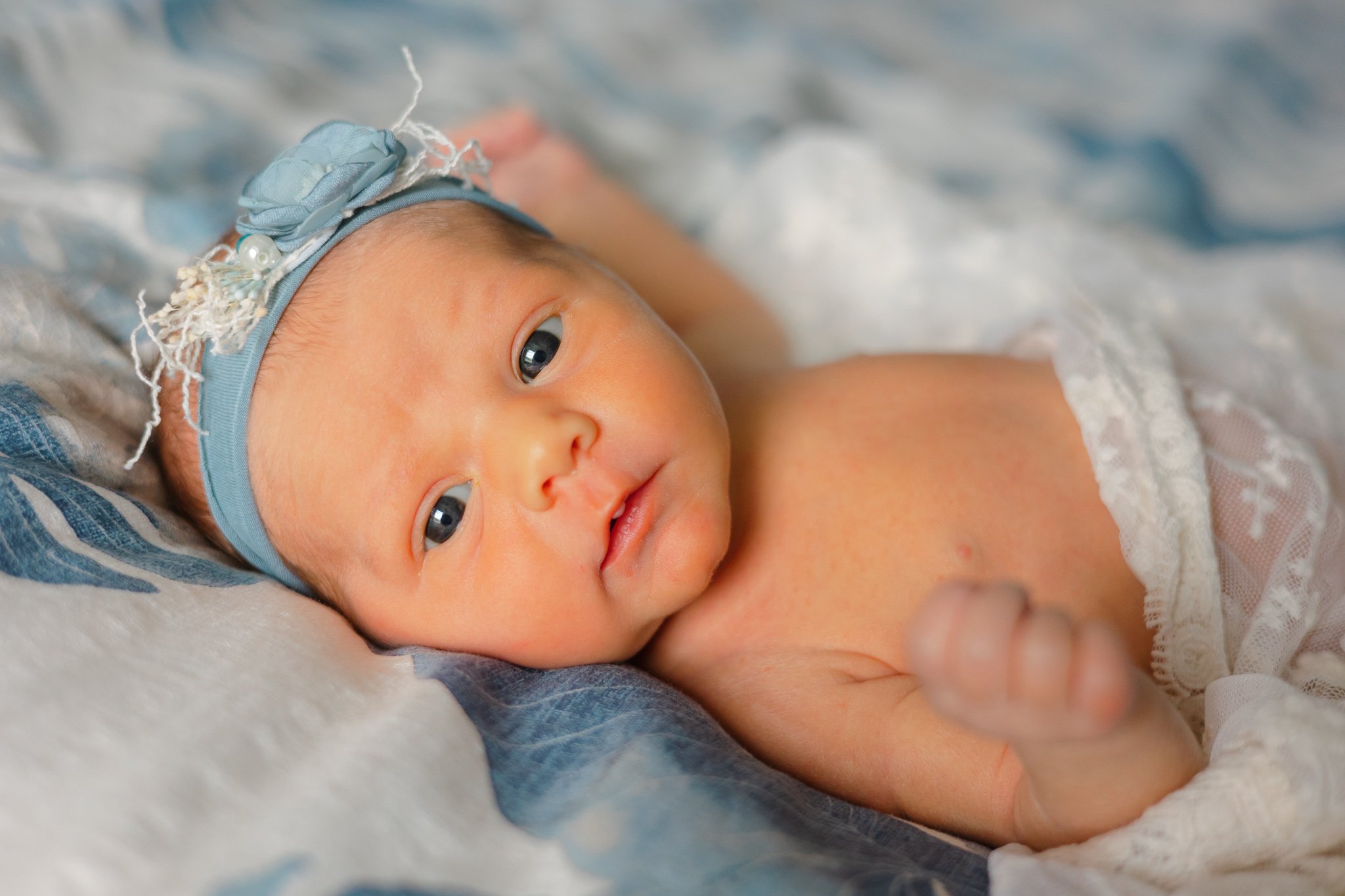 A newborn baby lays on a bed in a blue headband with eyes open southern sleep