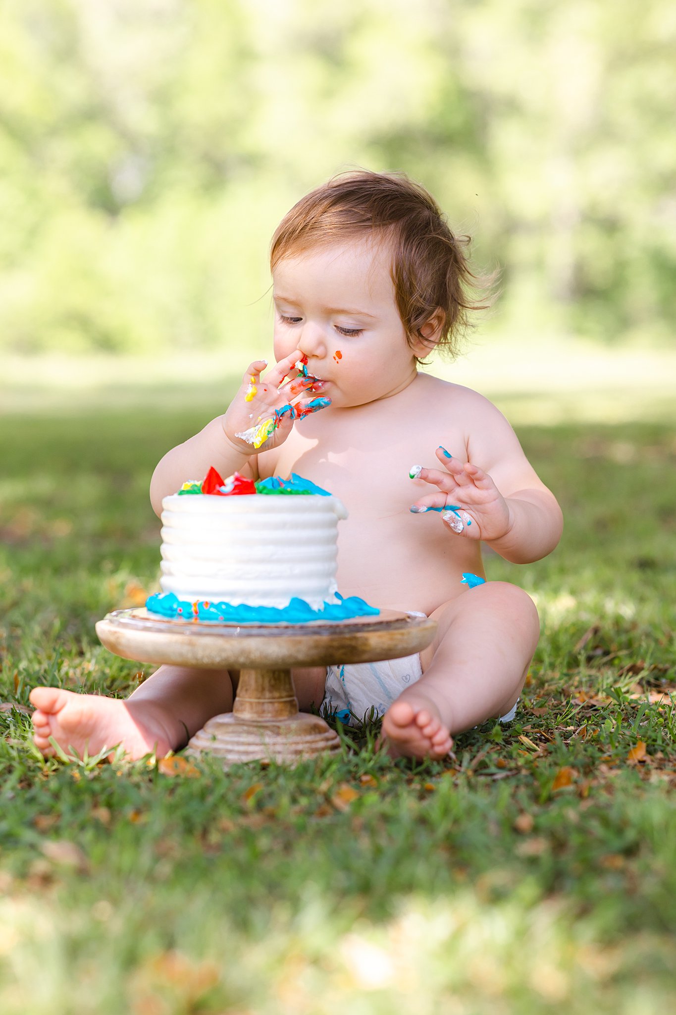 An infant baby licks frosting of its fingers in a cake smash on the grass in a park wearing a diaper before visiting daycares in savannah