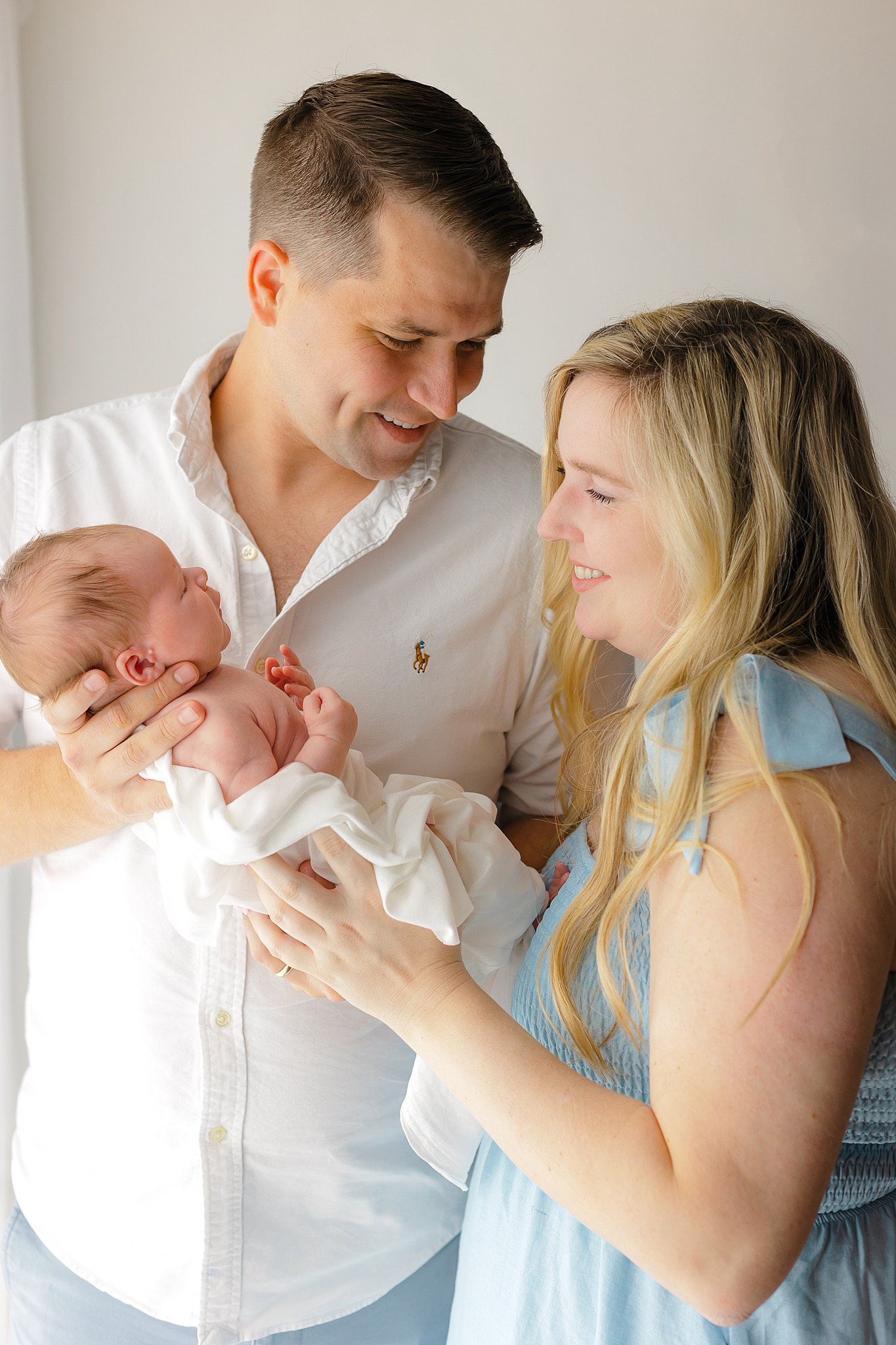 A mom smiles at her newborn baby in dad's hands as they stand together in a window