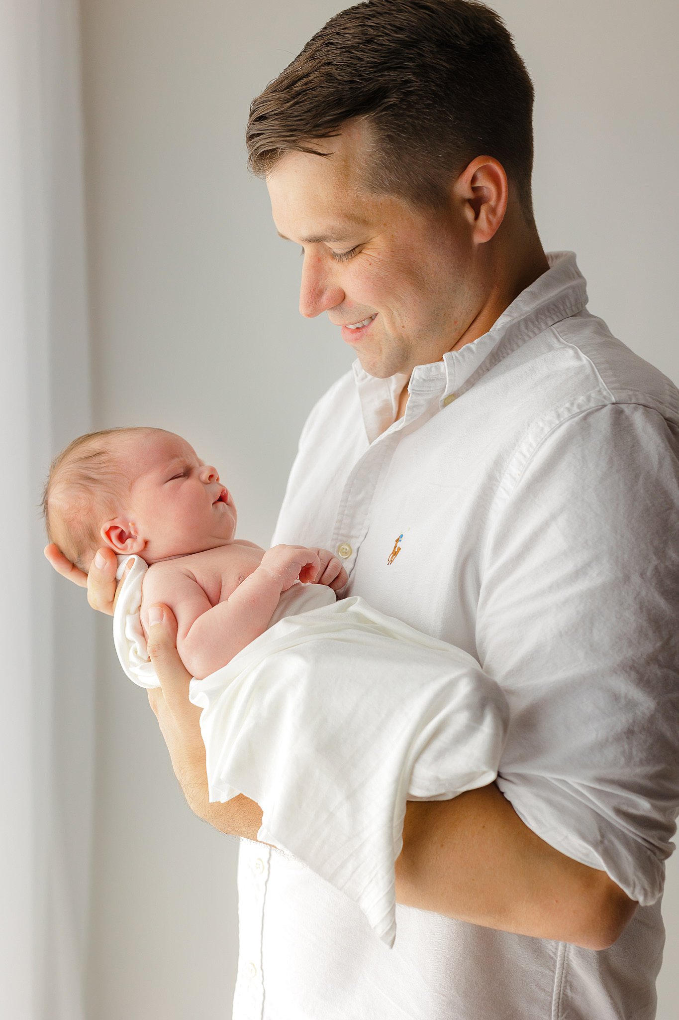 A new father in a white shirt smiles down to his sleeping newborn baby while standing in a window after visiting a savannah chiropractor