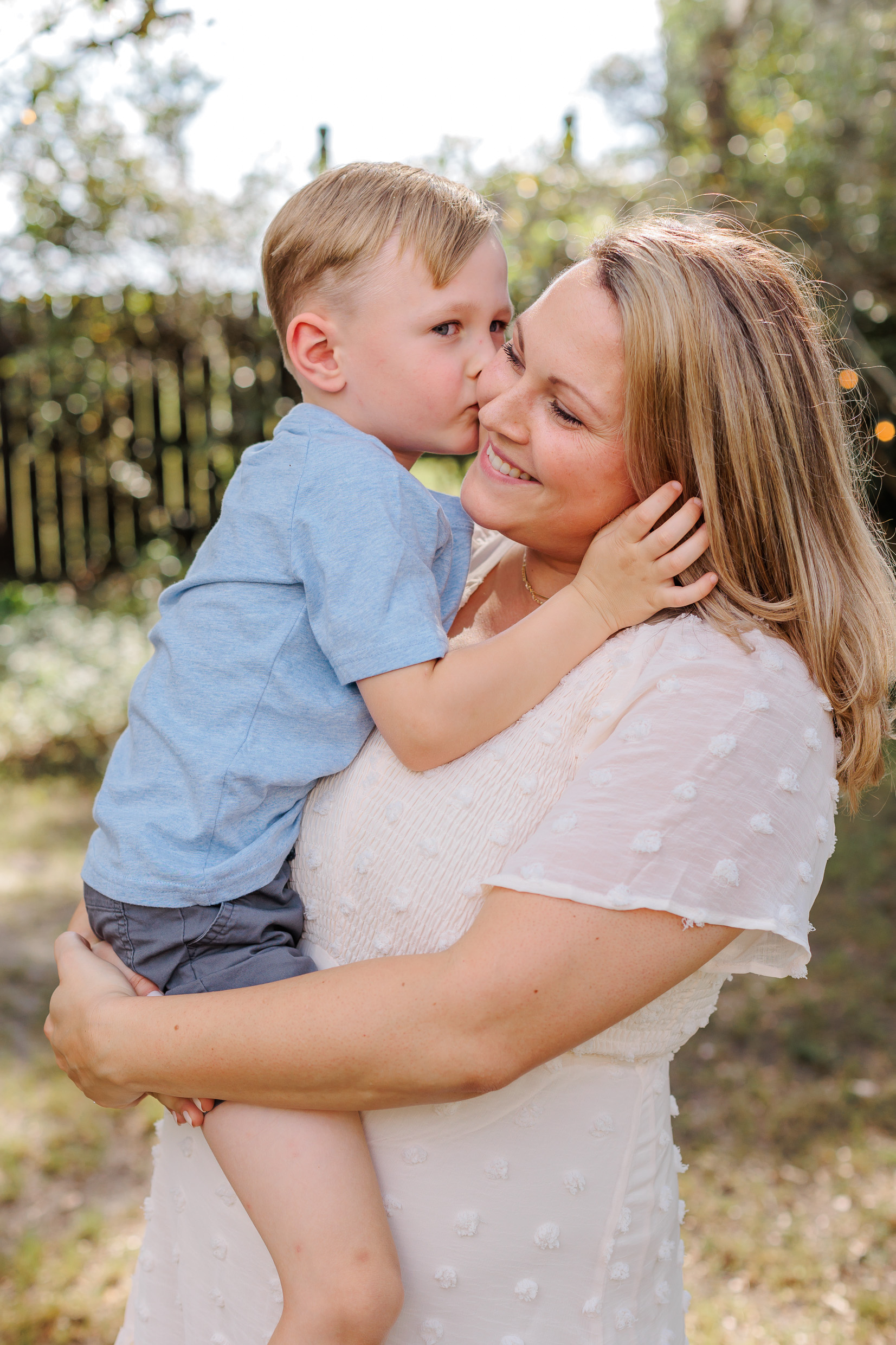 A mom in a white dress holds her young son on her hip as he kisses her in a blue shirt after visiting savannah preschools