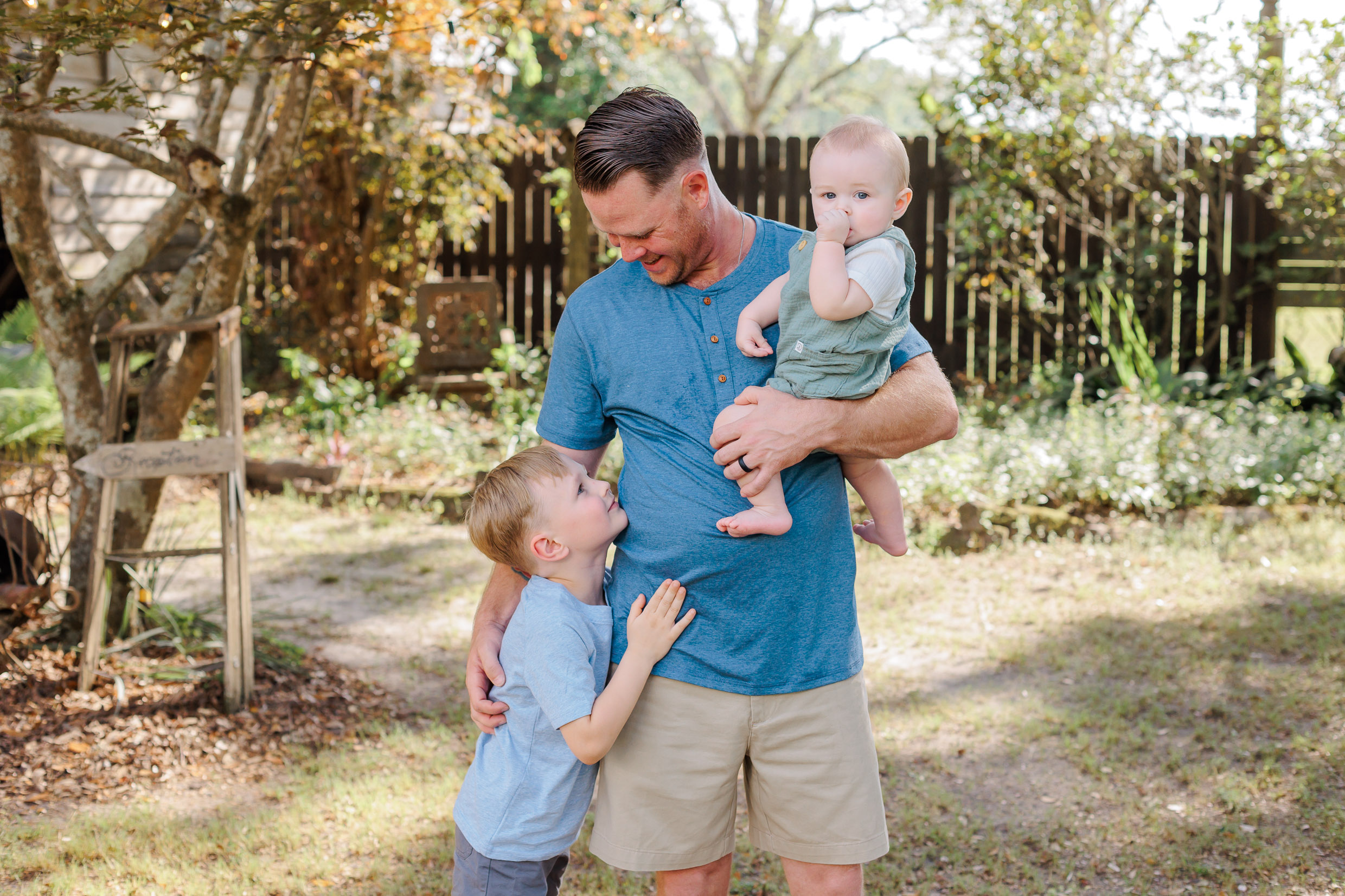 A father in a blue henley stands in a yard holding his toddler son in one arm and hugging onto his older son at his side thanks to savannah preschools