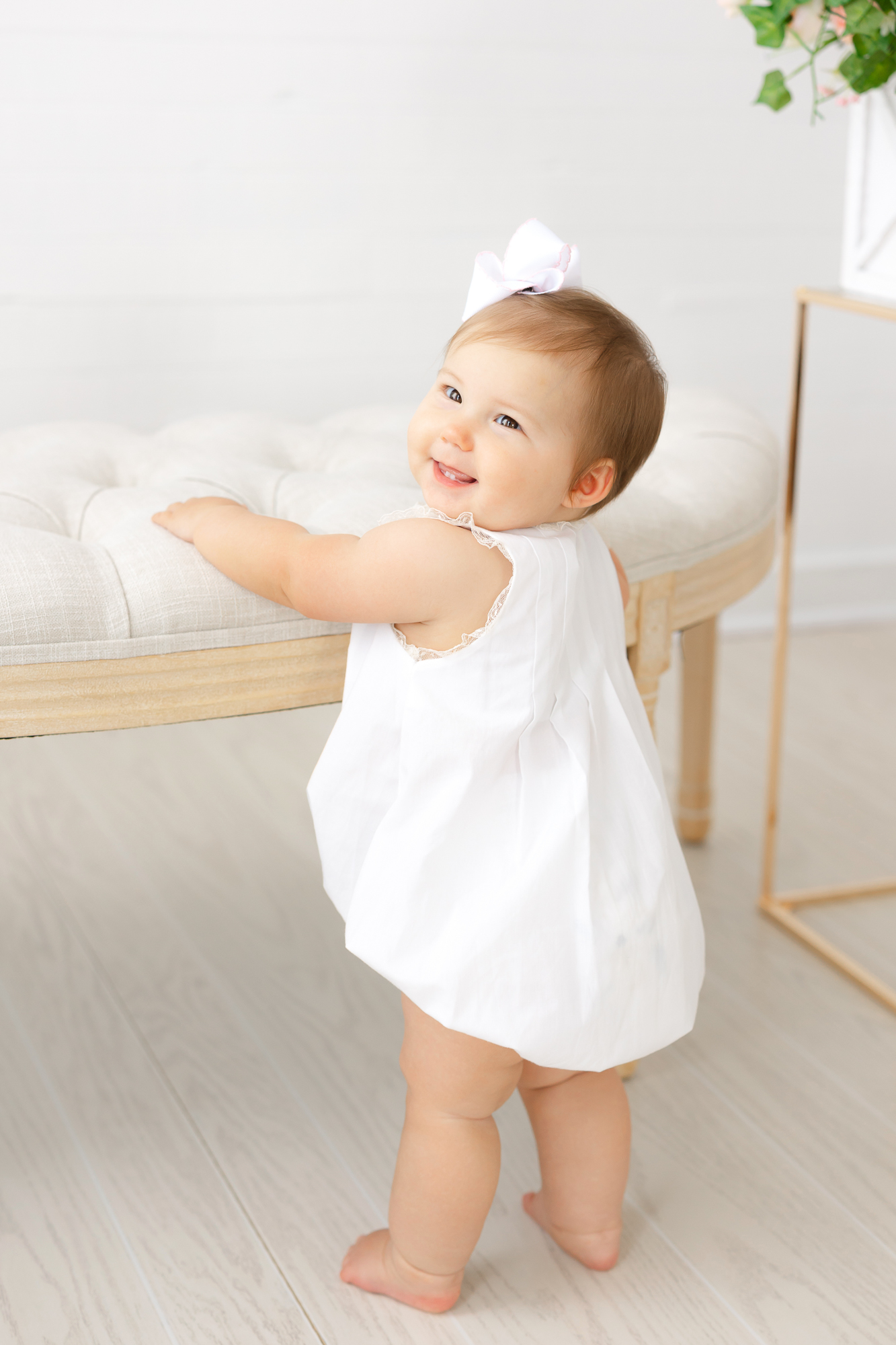Little girl wearing a white bubble outfit holding on to a small bench in Constance Calton Photography's studio in Springfield, Ga