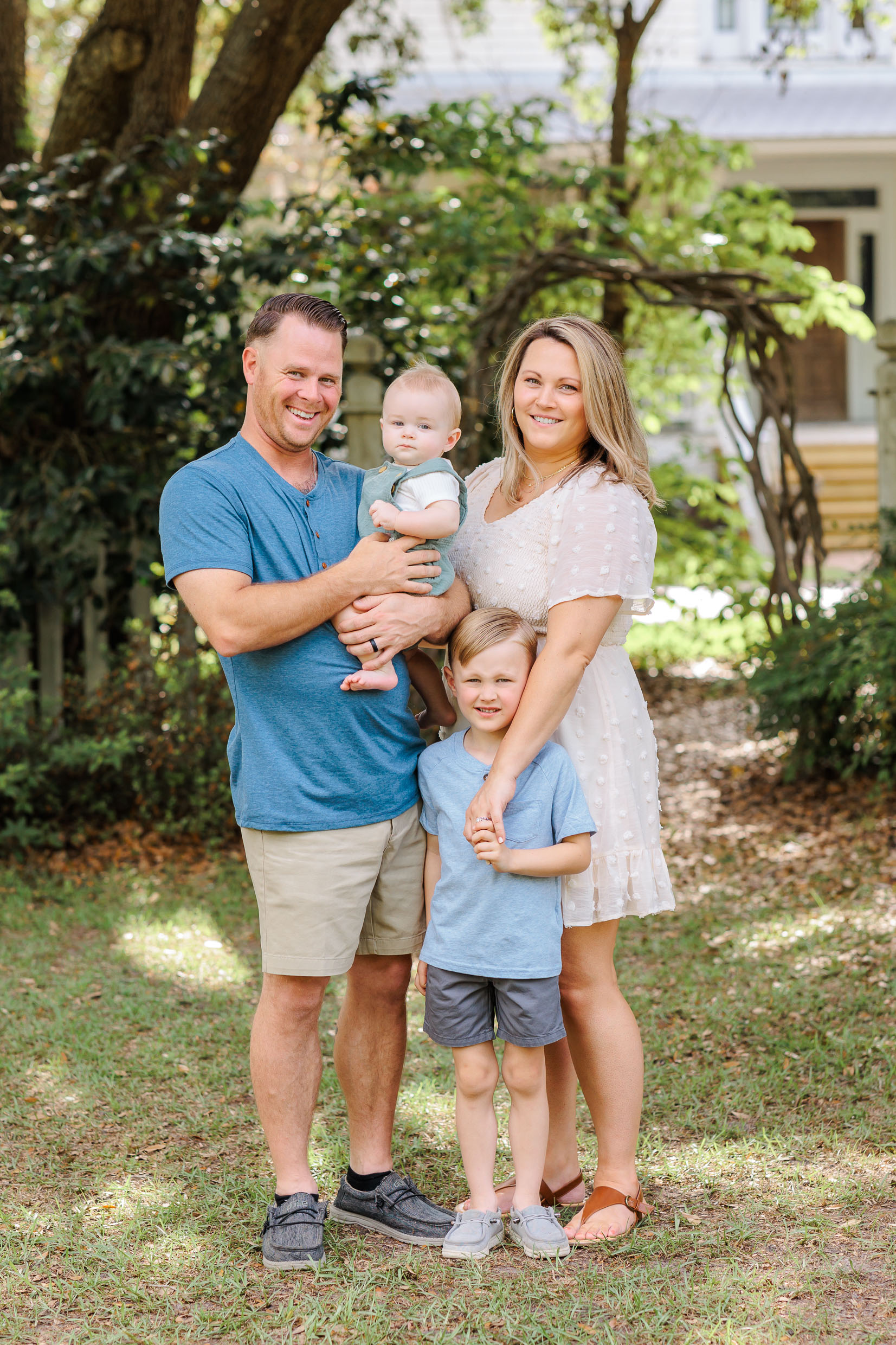 A mom and dad stand in a home garden with their infant son in dad's arms and young son holding mom's hand