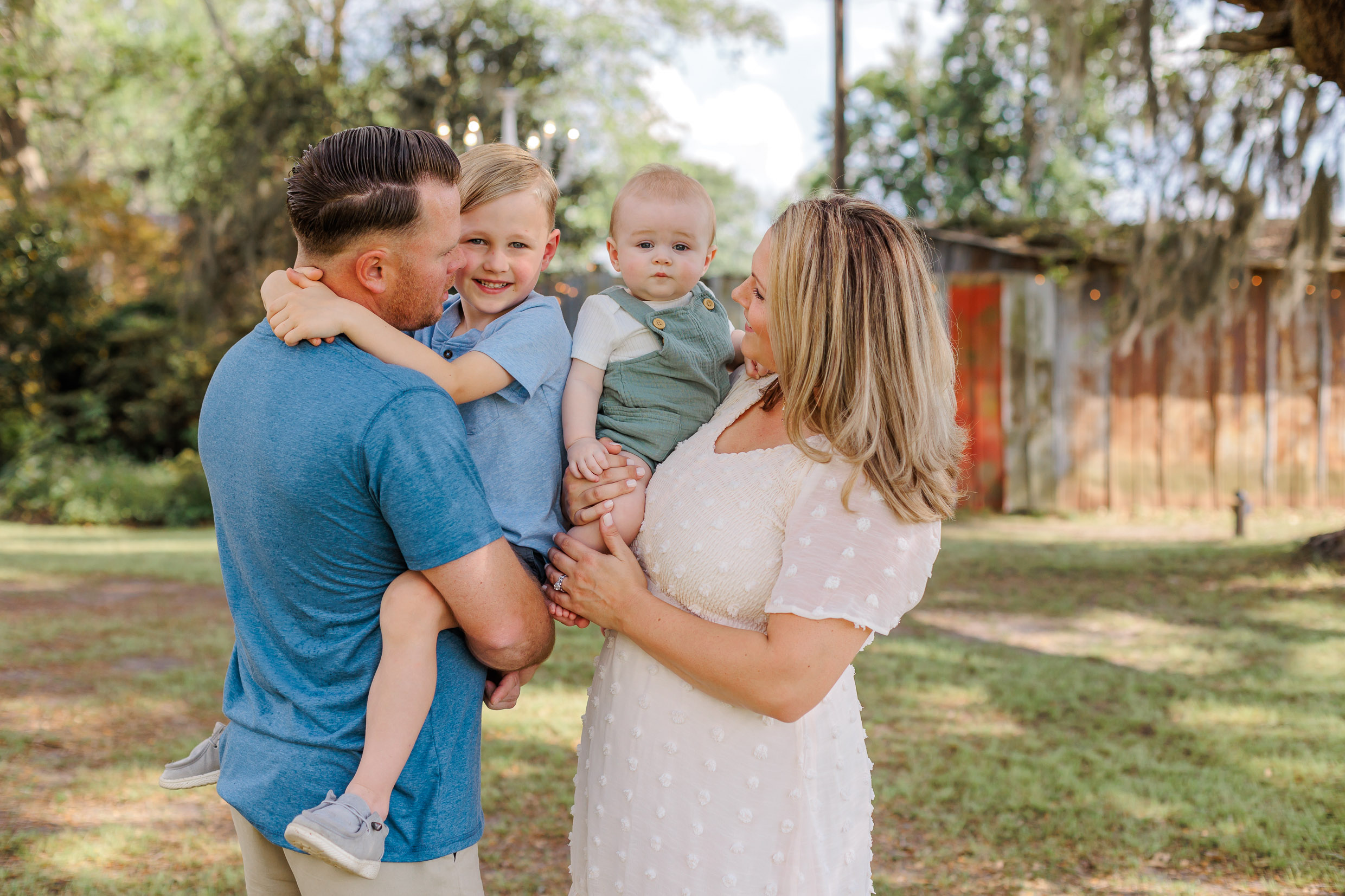 A mom and dad stand in a backyard holding their toddler son and young son in their arms after visiting preschools in pooler ga