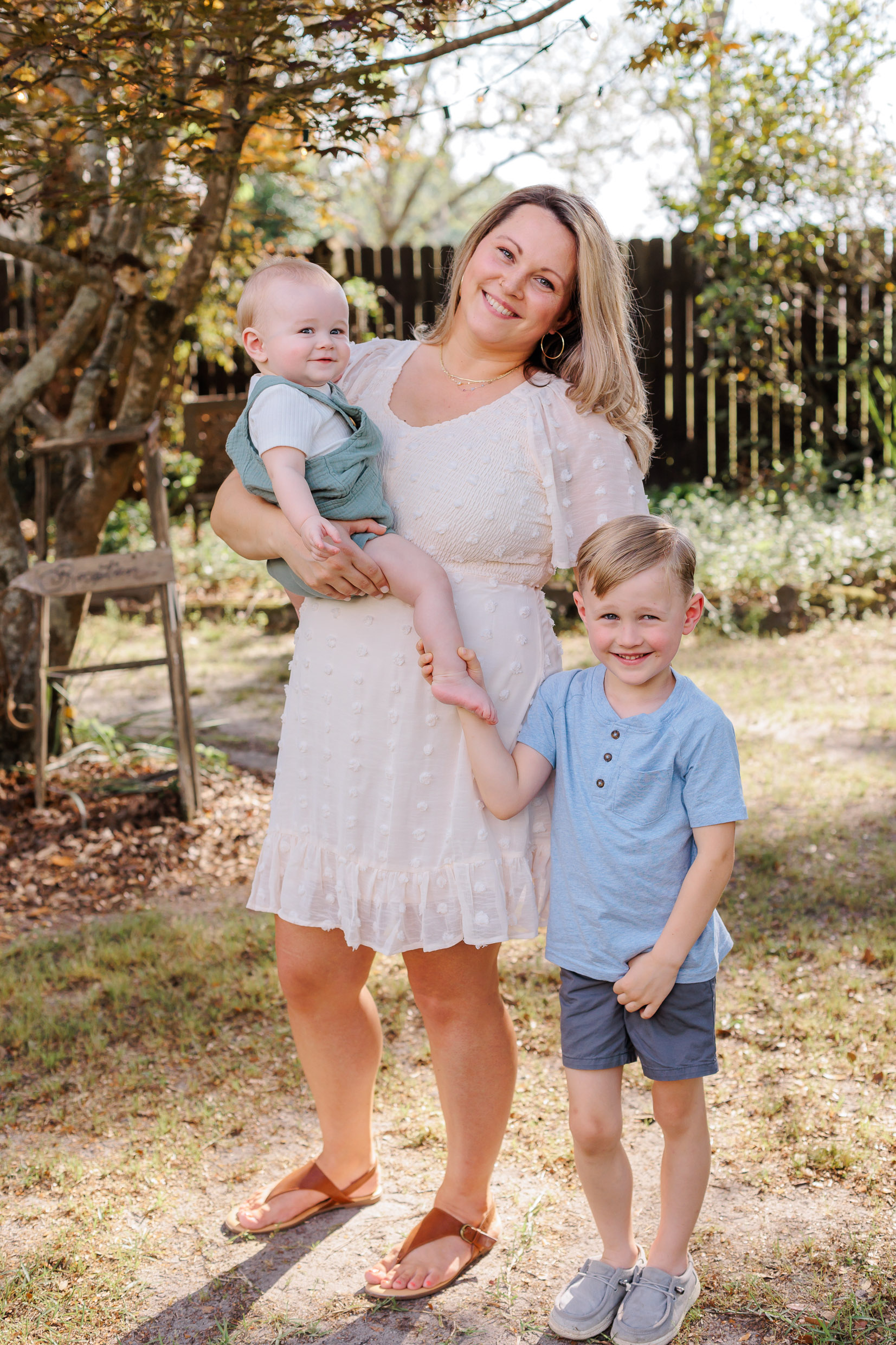 A mother smiles while standing in a garden yard with her toddler on her hip and young son at her side holding the toddler's foot after attending preschools in pooler ga