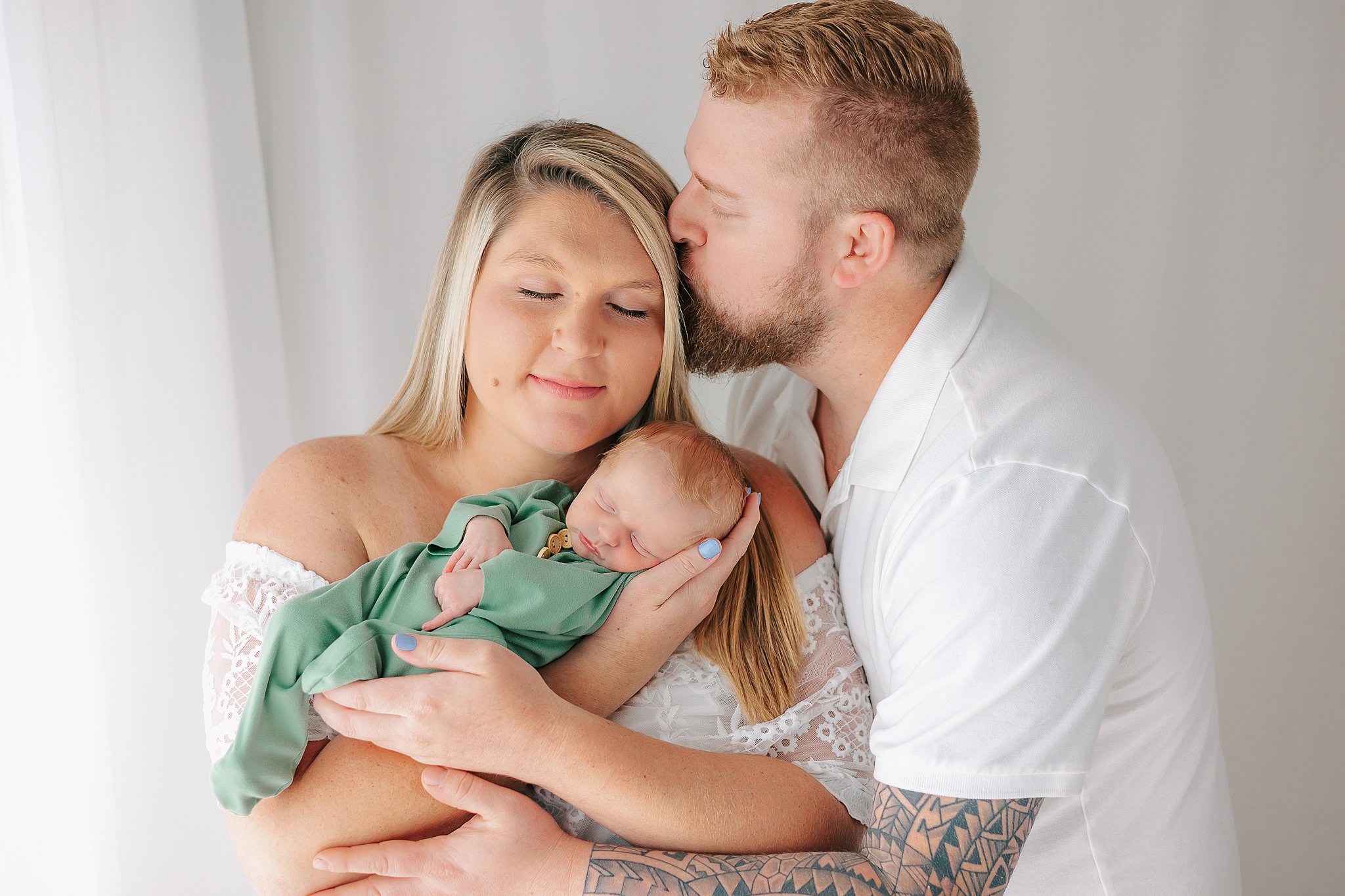 A husband hugs and kisses his wife as she holds their sleeping newborn baby