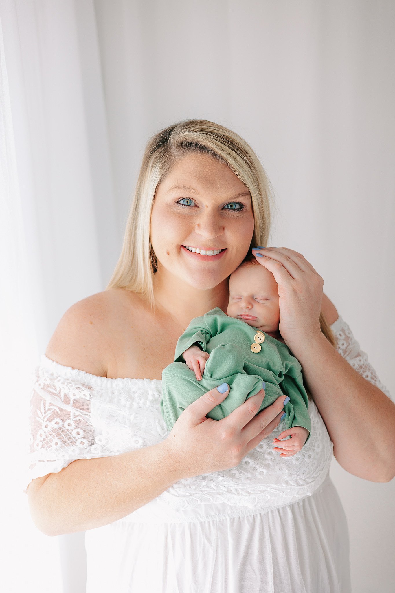 A happy mom stands in a window in a white lace dress while cradling her sleeping newborn baby against her chest after getting help from a savannah obgyn