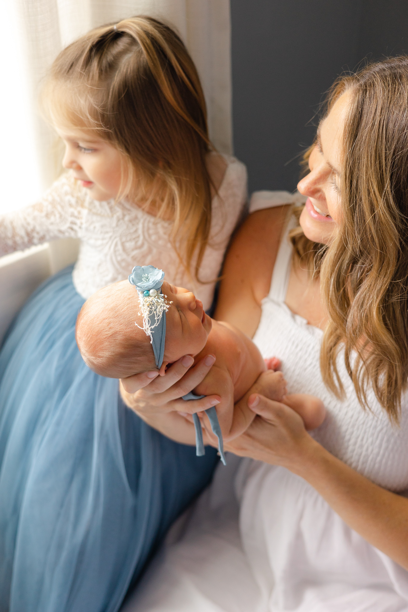 A mom in a white dress sits on the floor showing her newborn baby to her toddler sister