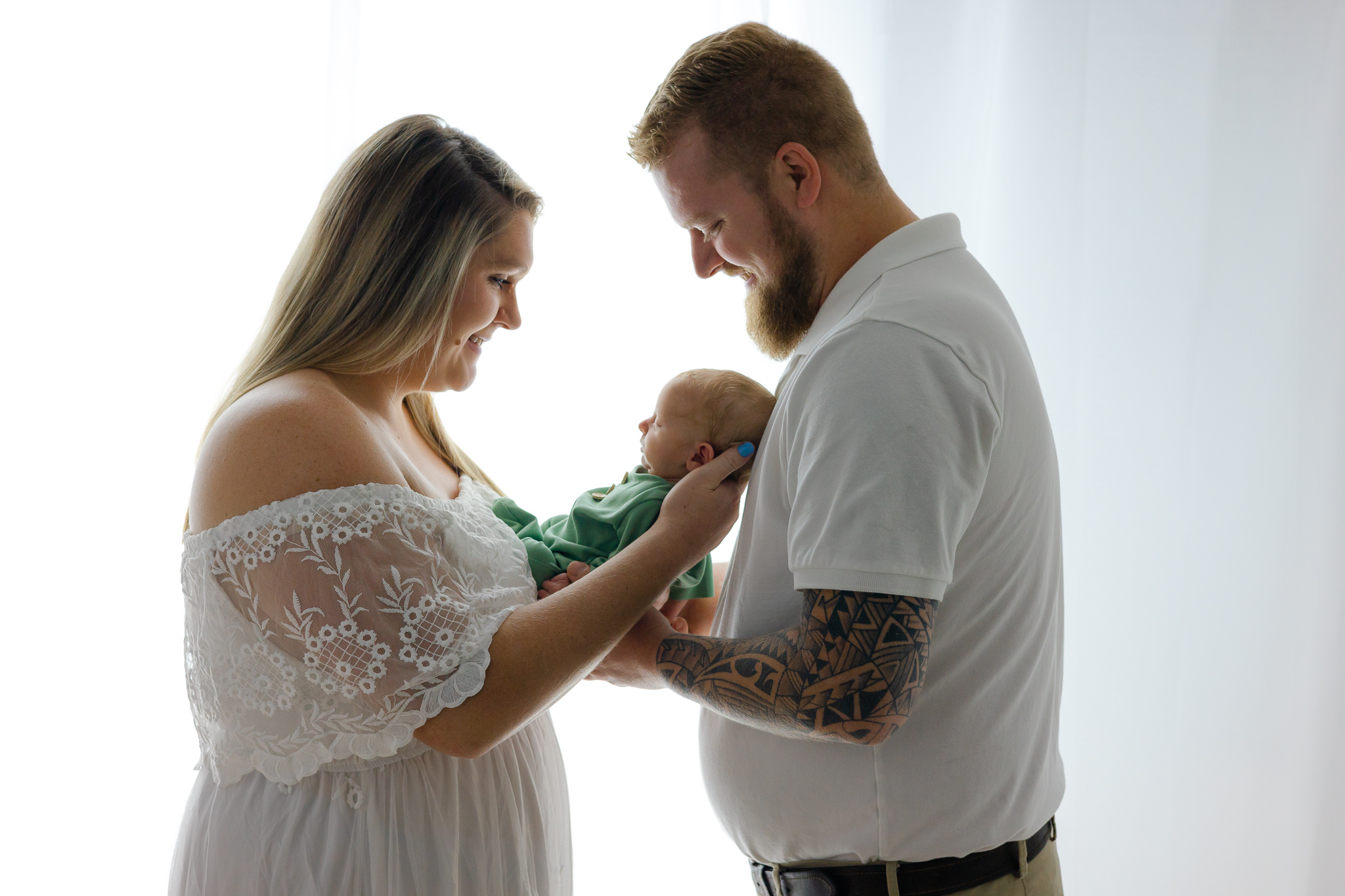 A mom and dad both in white stand in a studio in a window smiling down to their newborn baby in their hands