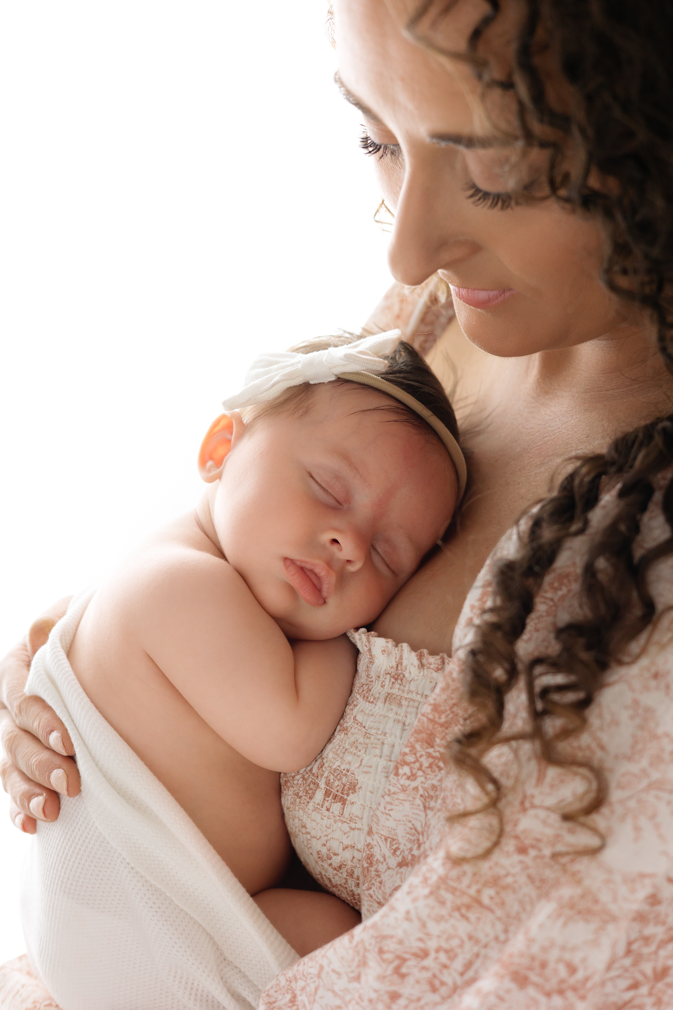 A mom dressed in pink stand in a studio in a window smiling down to her newborn baby on her chest sleeping