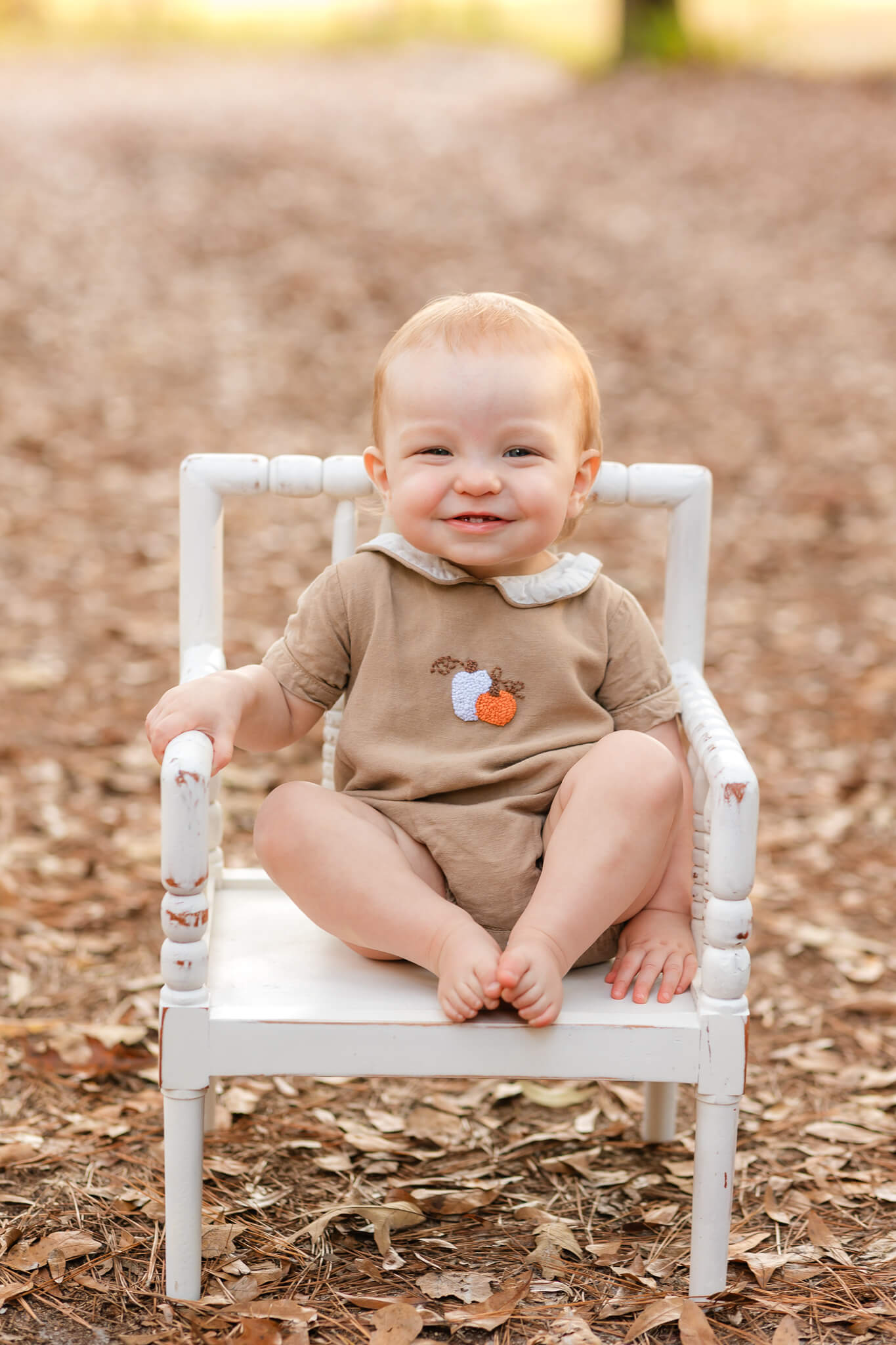 One year old boy wearing a brown fall outfit with a pumpkin. He is sitting in a while chair outside with brown leaves surrounding him. Pumpkin patches in Savannah, Ga