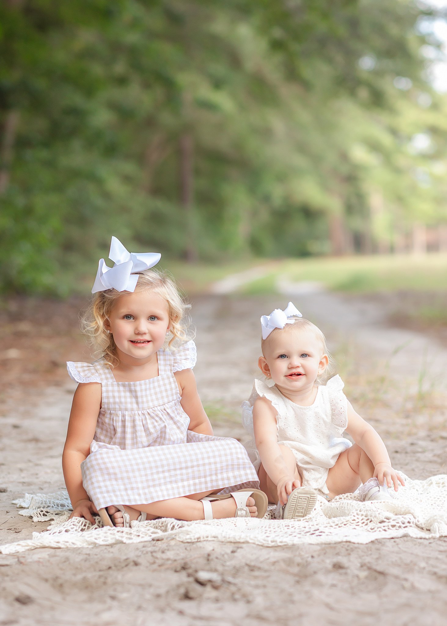 Toddler sisters in pink dresses and white bows sit on a lace blanket in a sandy trail in a park after meeting Babysitters in Savannah, GA