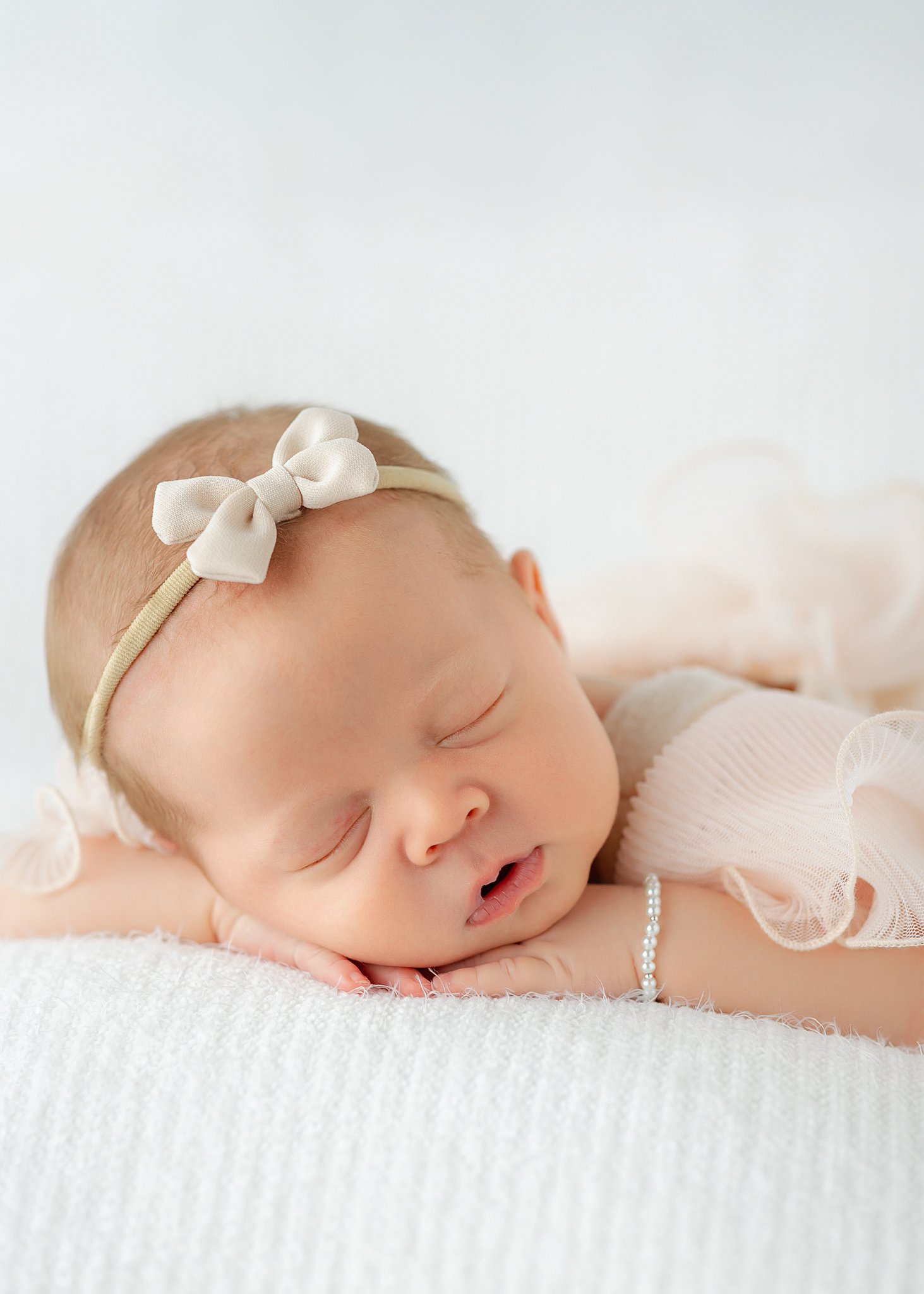 A cute newborn bay girl sleeps on a white bed with head on her hands in a tiny bow headband