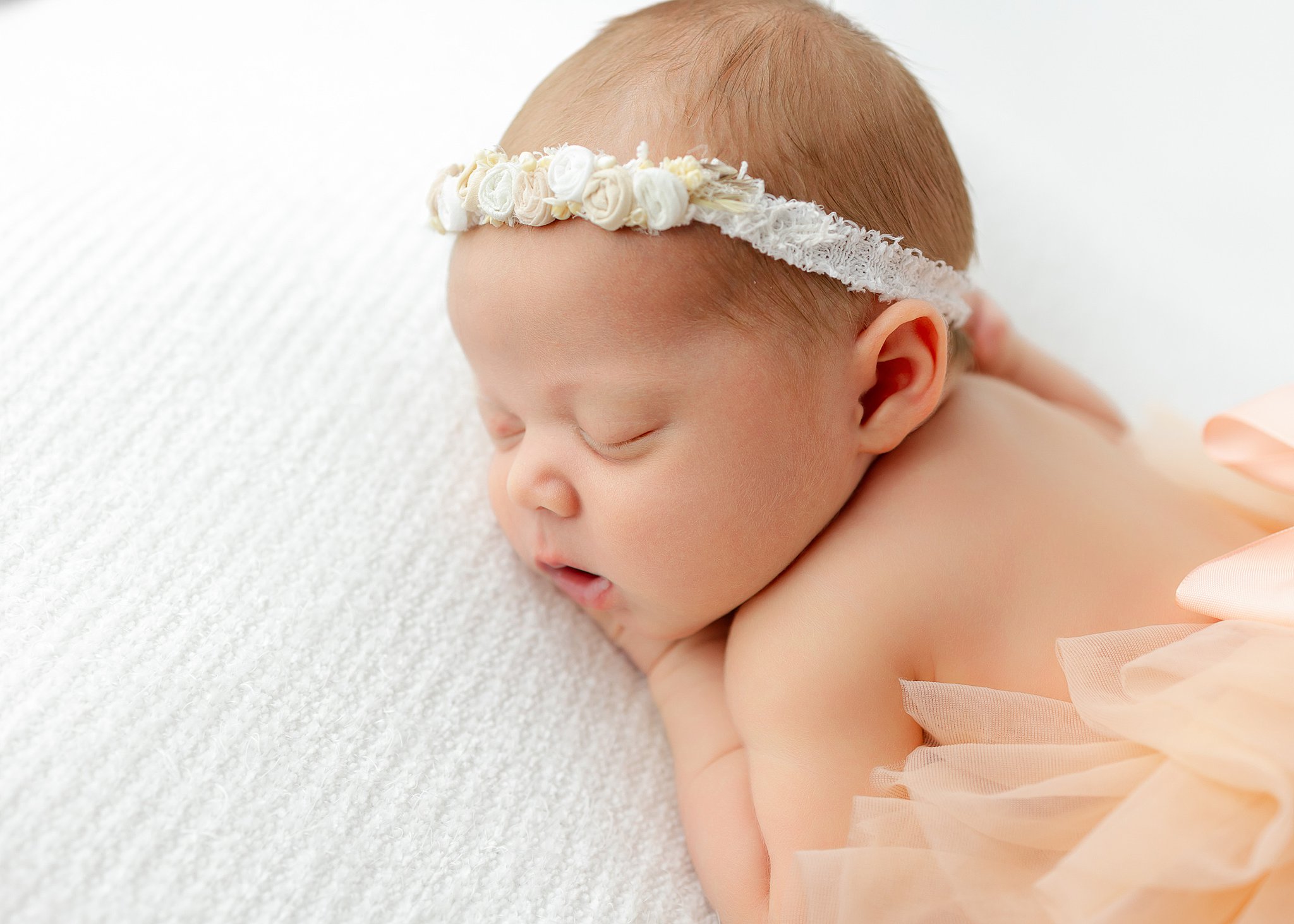 A newborn baby in a floral headband sleeps on her belly on a white bed after meeting Lactation Consultants in Savannah, GA