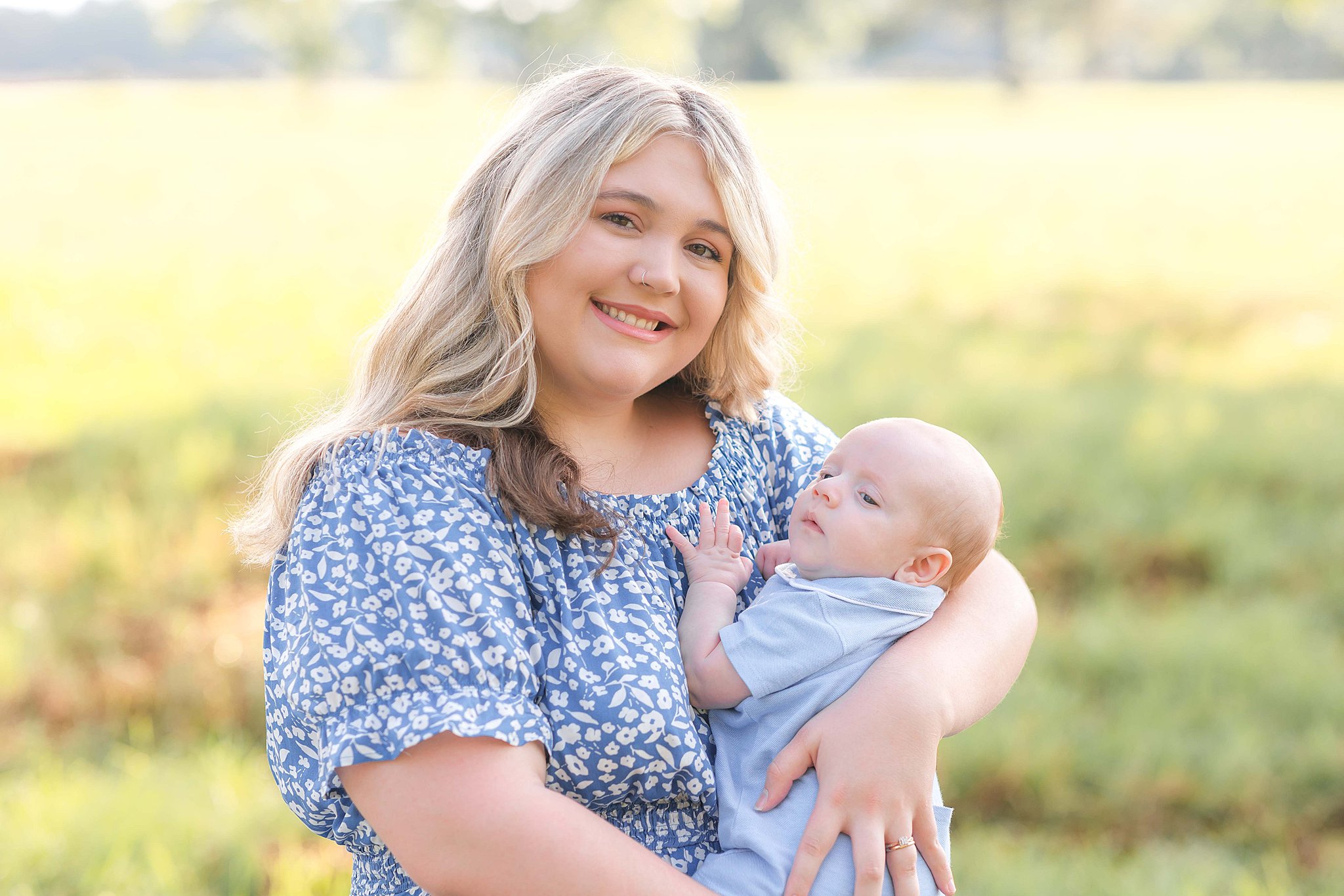 A happy mother in a blue floral dress stands in a field at sunset holding her baby against her chest