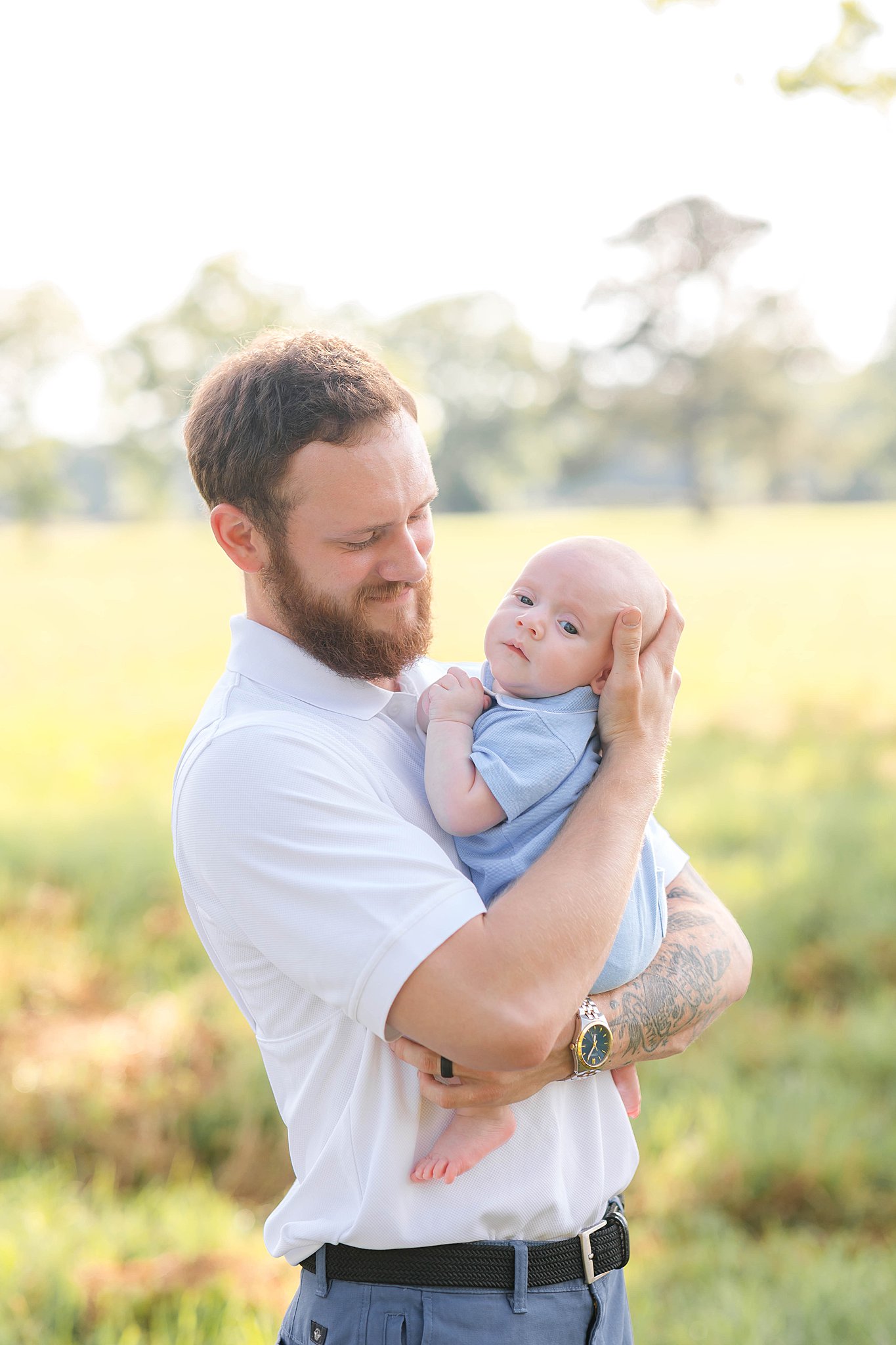 A happy father cradles his baby in a blue onesie against his chest while standing in an open field after visiting the bunny hive savannah