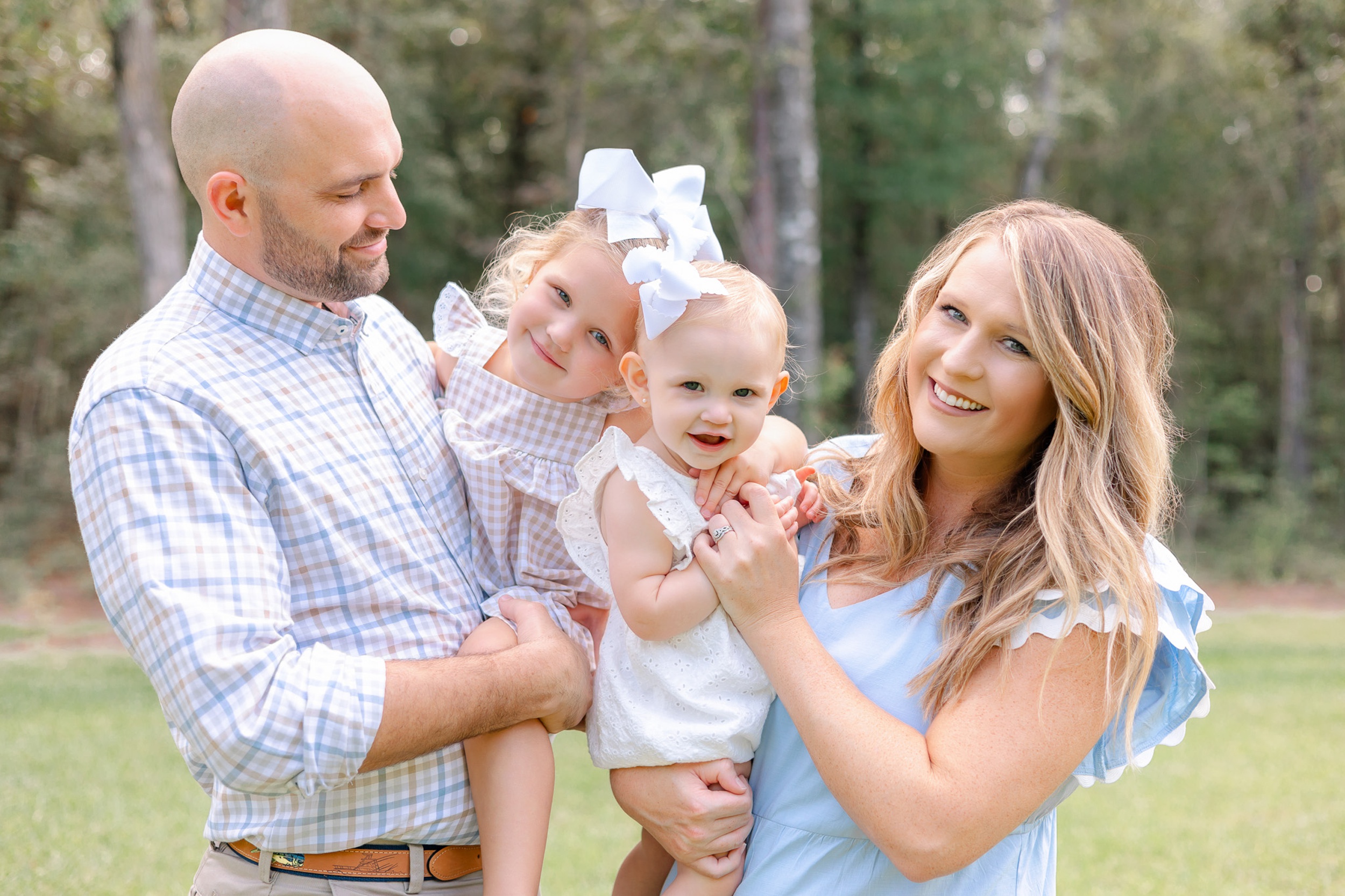 Happy mom and dad hold their toddler daughters on their hips while in a park lawn in dresses