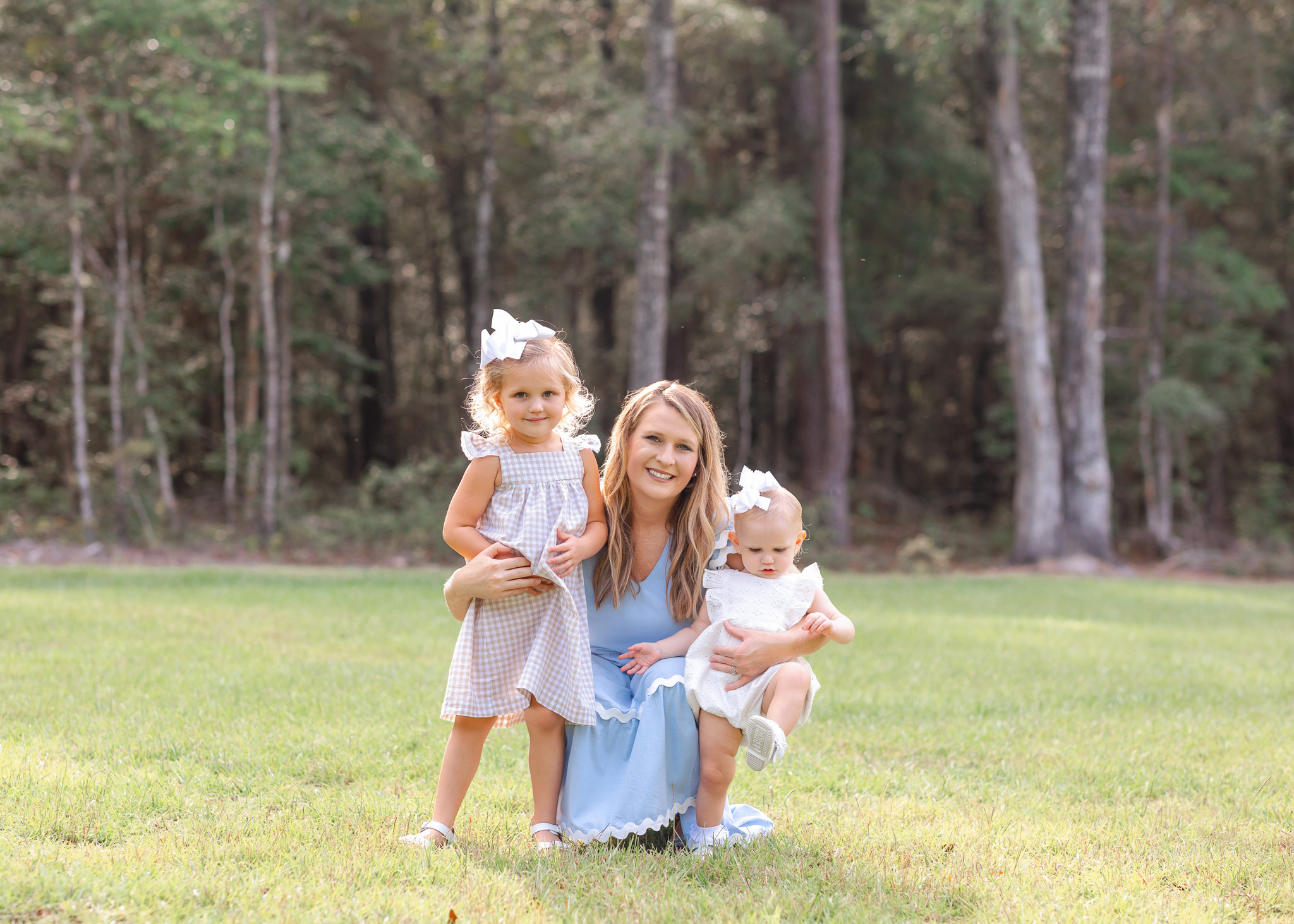 A happy mom in a blue dress kneels in a park lawn with her two toddler daughters after some parenting classes in Savannah, GA